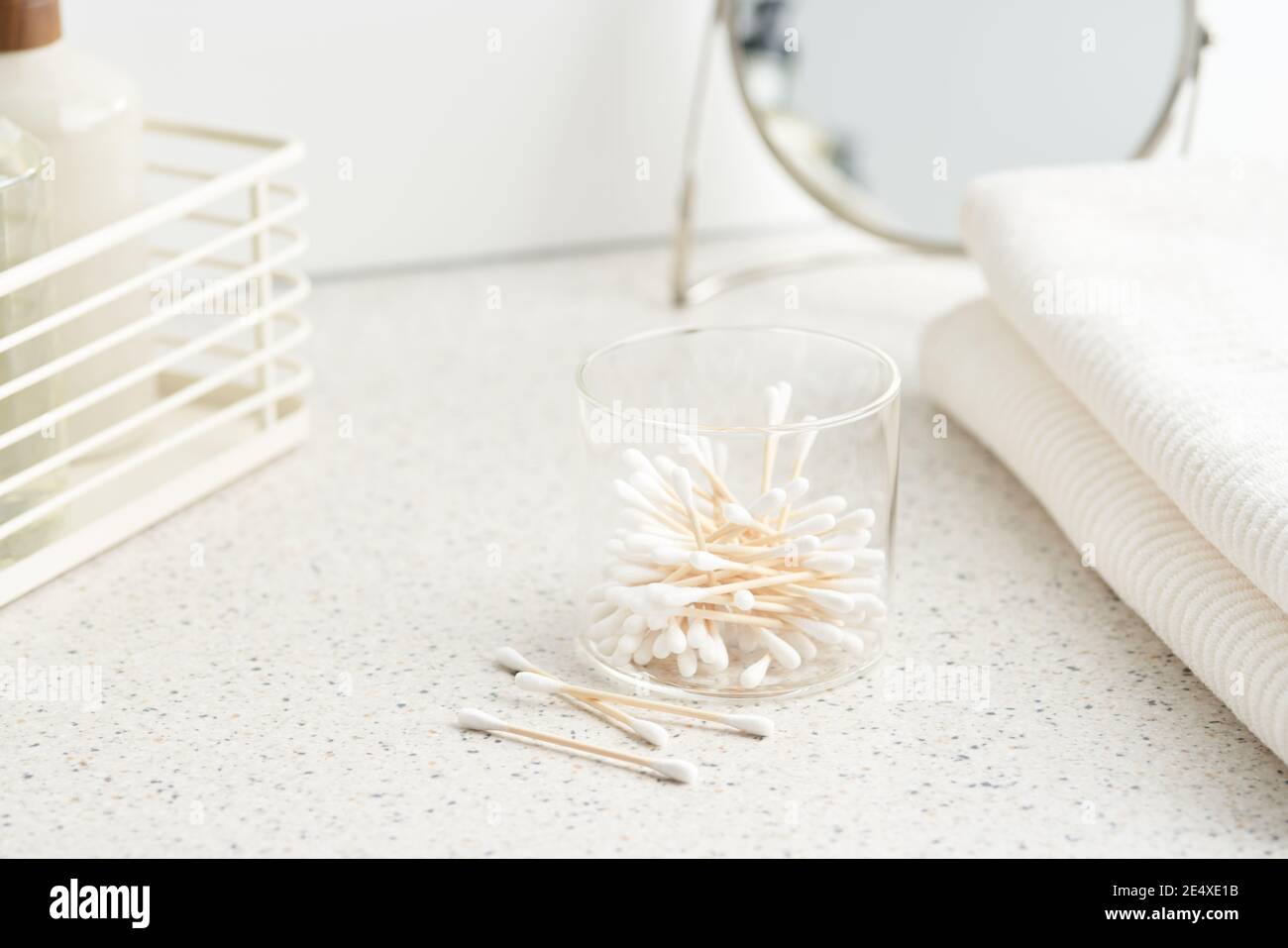 Contenitore di vetro con tamponi di cotone di bambù posizionati vicino alla pila di asciugamani al mattino in bagno Foto Stock