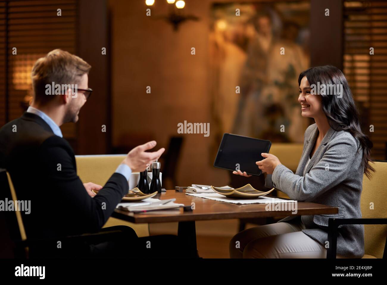 Gioiosa bruna signora che condivide un tavolo da pranzo con il giovane e mostrargli un tablet con schermo vuoto Foto Stock