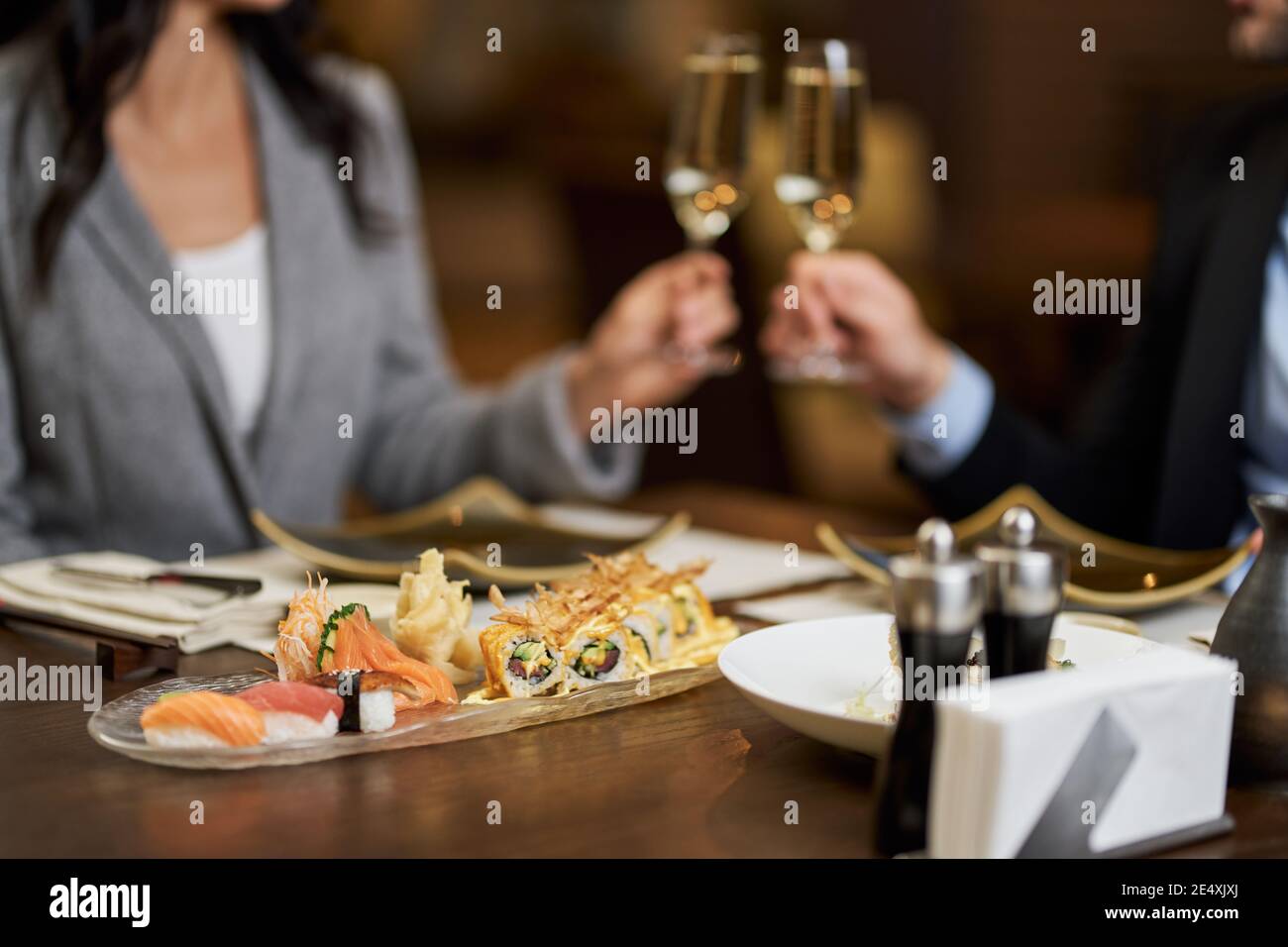 Foto a fuoco selettivo di delizioso sushi su un piatto con coppia sfocata che graffia bicchieri di champagne sullo sfondo Foto Stock