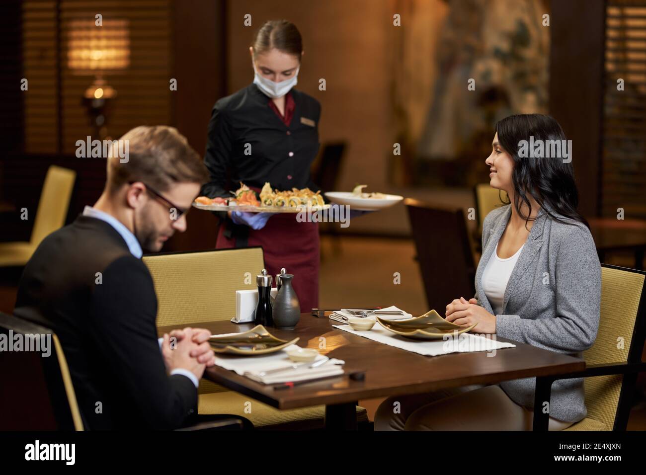 Due giovani che hanno una data al tavolo del ristorante mentre cameriera ben addestrata in maschera portando la loro cena Foto Stock