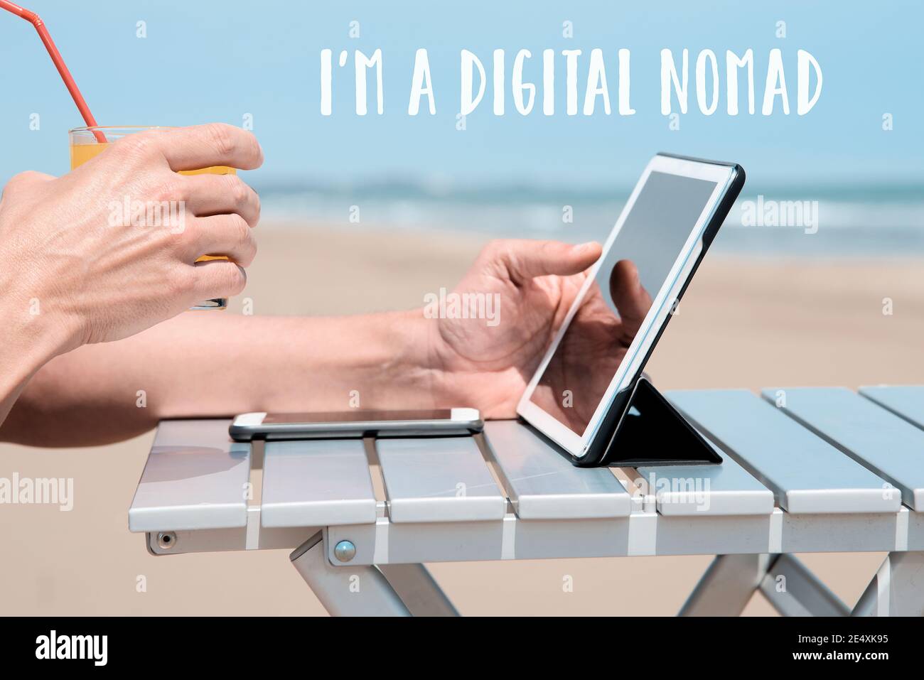 closeup di un giovane caucasico sulla spiaggia, seduto a un tavolo all'aperto, utilizzando un tablet digitale, mentre è una rinfrescante bevanda arancione, e. Foto Stock