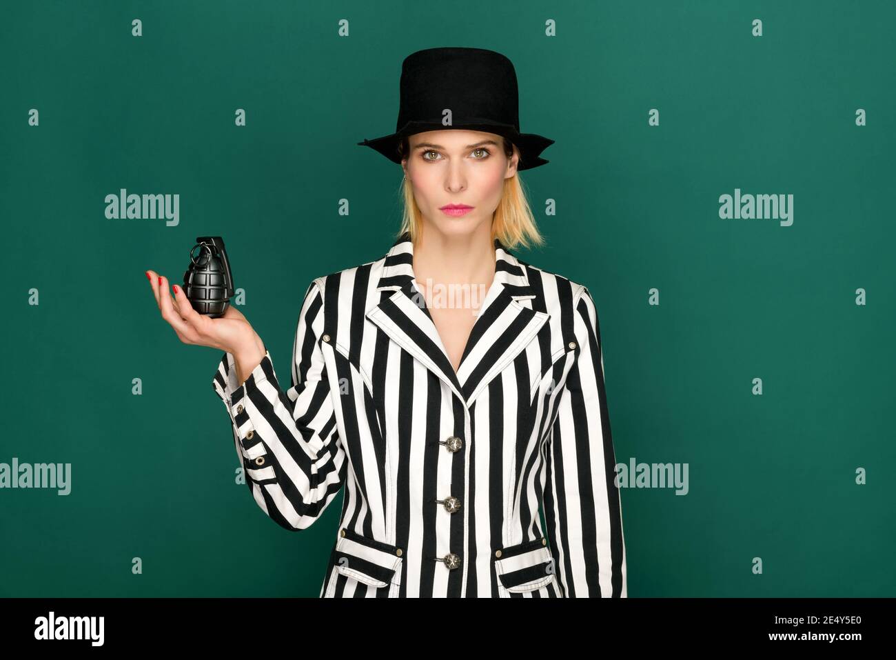Modello biondo elegante e sicuro con giacca a righe in bianco e nero e cappello che tiene la granata nera su sfondo verde Foto Stock
