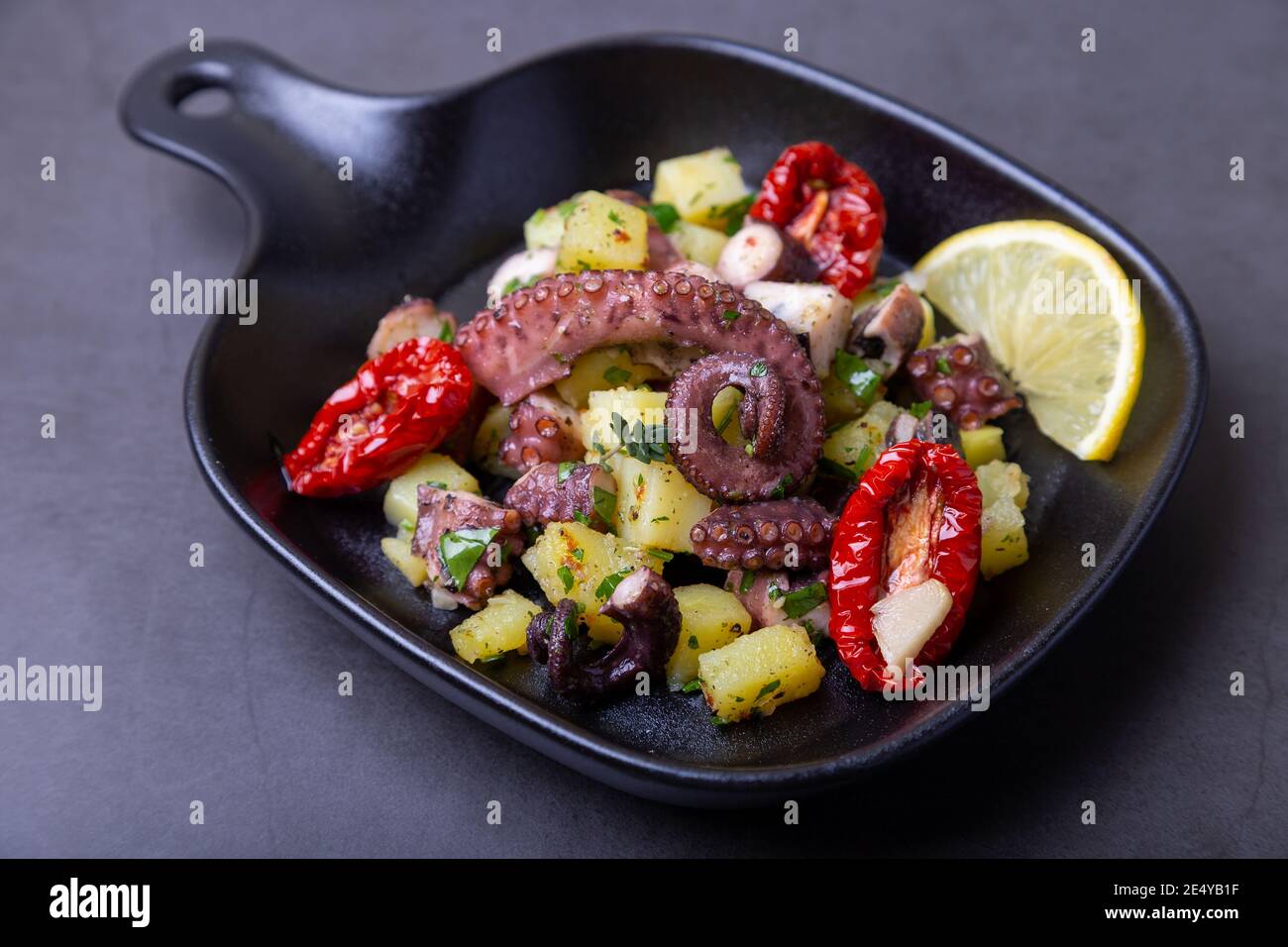 Insalata tiepida con polpo, patate, pomodori secchi, aglio e limone in una piccola padella nera. Primo piano, sfondo nero. Foto Stock