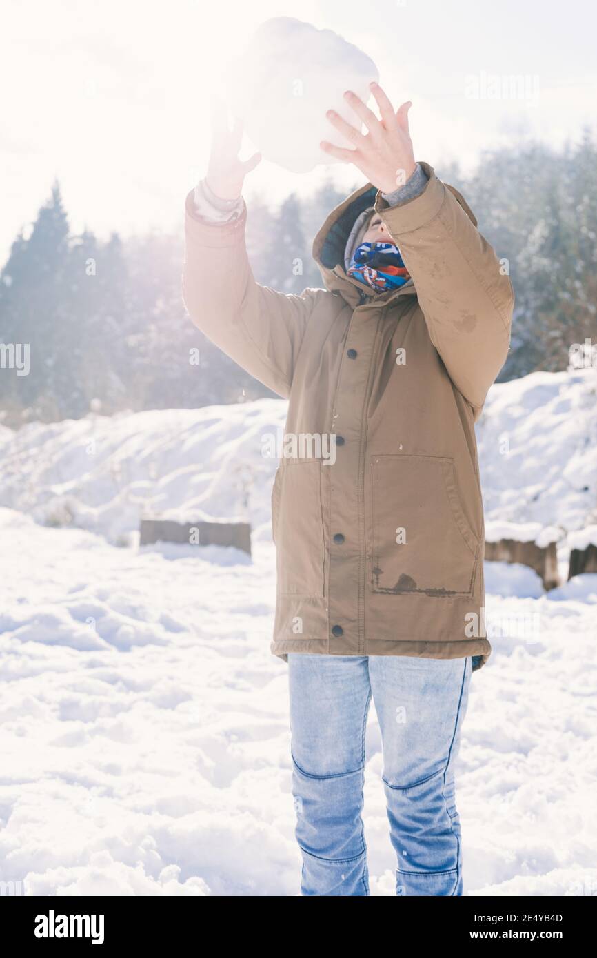 Messa a fuoco superficiale di un ragazzo in una giacca marrone cattura una grande palla di neve Foto Stock