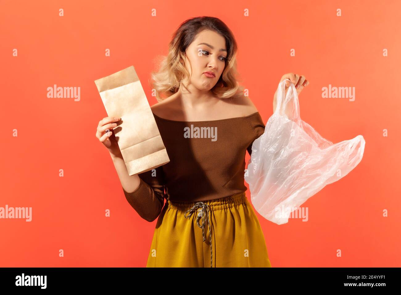 Giovane donna che tiene in mani di carta e sacchetti di plastica, preferendo prodotti ecologici, campagna contro la plastica. Studio interno isolato su arancione b Foto Stock