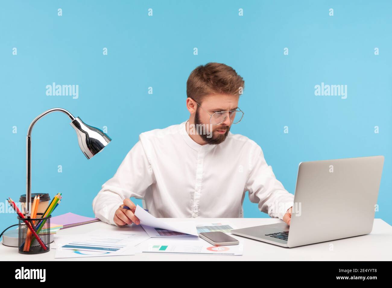 Analista uomo concentrato in occhiali e camicia bianca che controlla i dati sui grafici, inserendo i risultati al programma del laptop, seduto sul posto di lavoro. Prigioniero interno Foto Stock