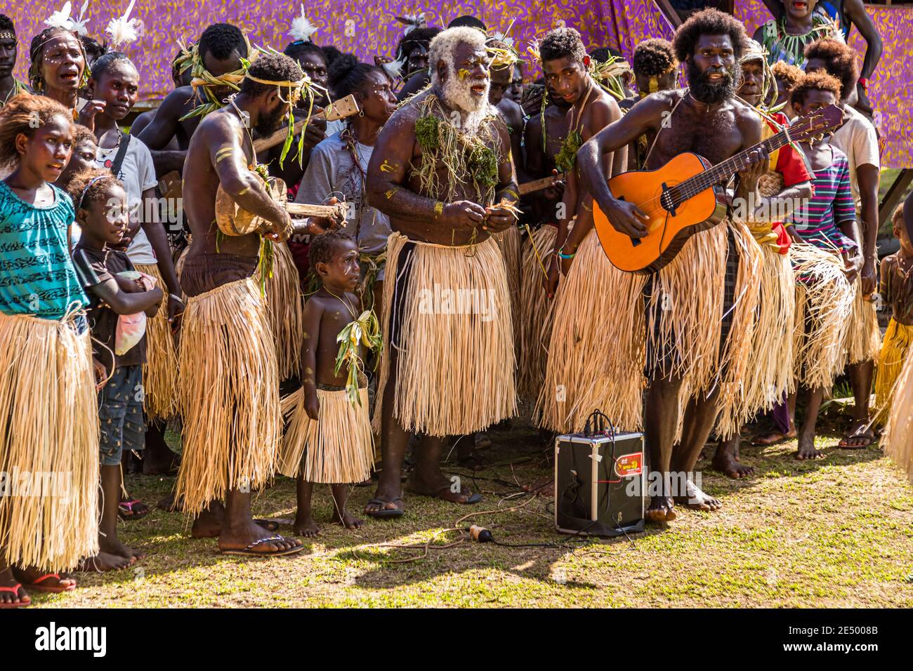 Chitarristi e suonatori di ukulele accompagnano un uomo anziano con la barba bianca e il muschio drappeggiato sul petto durante un festival del villaggio Sing-Sing a Bougainville, Papua Nuova Guinea, circondato da spettatori davanti a un padiglione tenda viola. Foto Stock