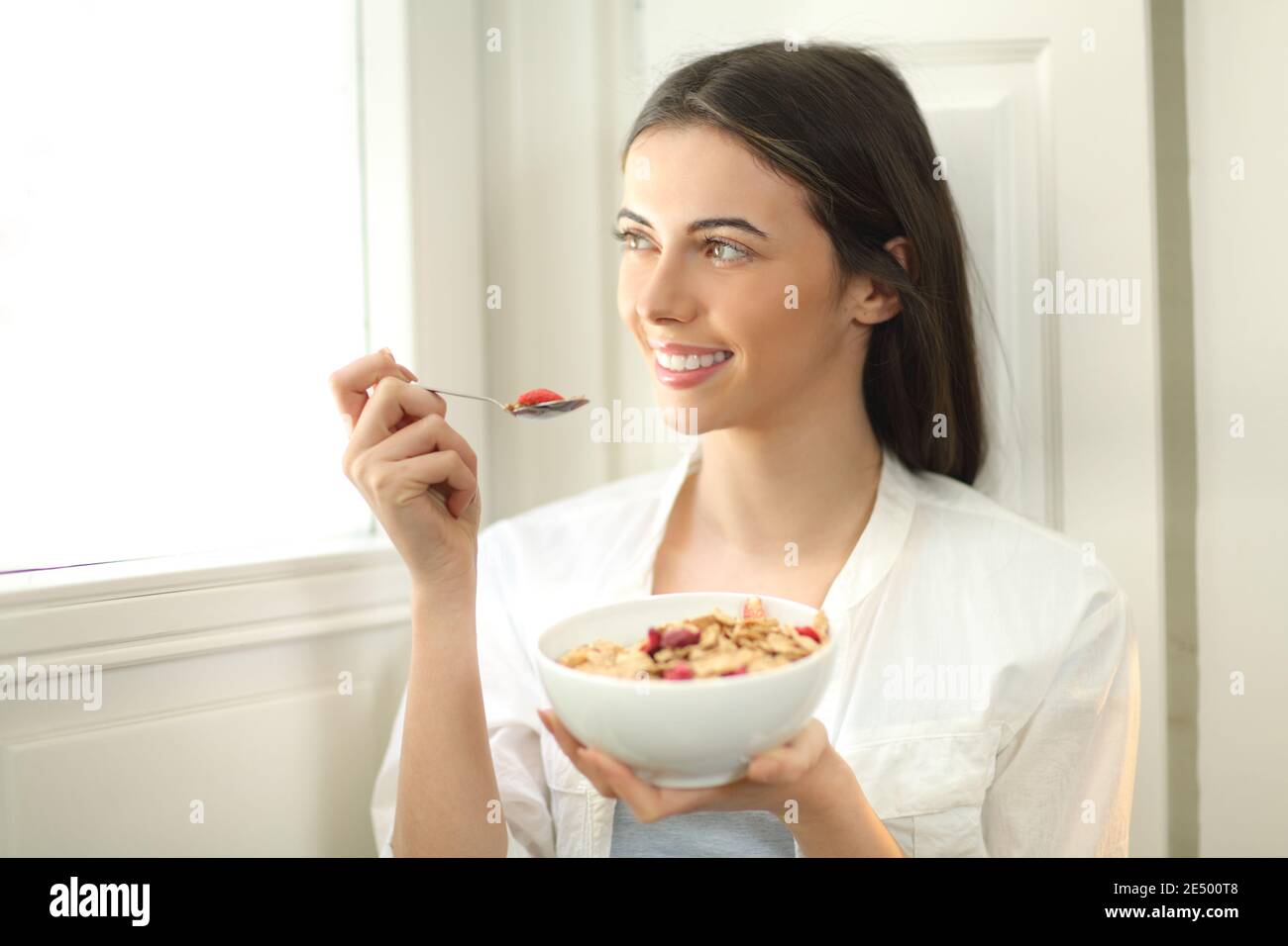 Buona donna che mangia cereali a colazione la mattina guardando attraverso una finestra a casa Foto Stock