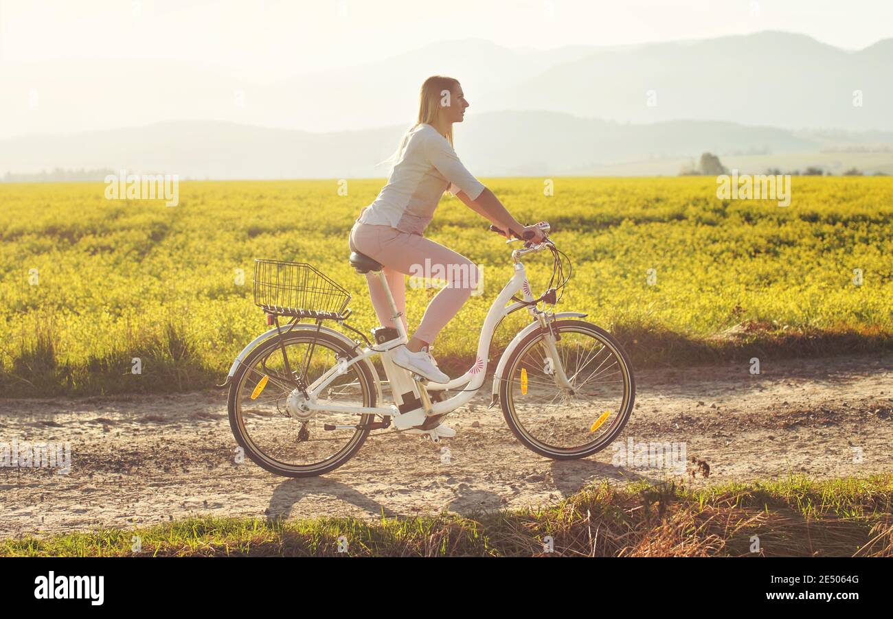 La giovane donna corre in bicicletta elettrica su polverosa strada di campagna, vista da lato, campo di retroilluminazione del sole con sfondo di fiori gialli Foto Stock