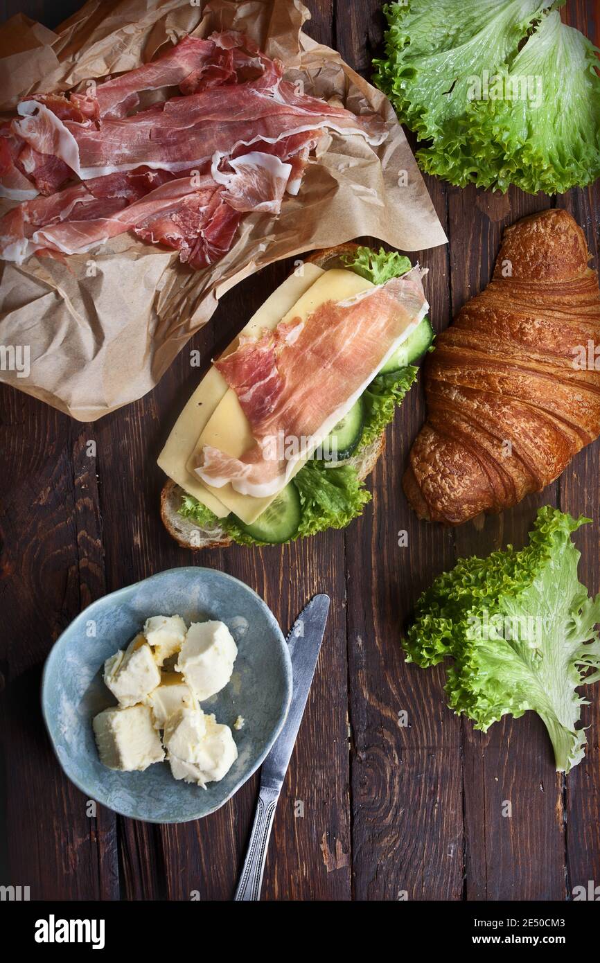 cucina rustica tavolo in legno con panino croissant, fette di carne di jamon, formaggi stagionati, foglie di lattuga verde, cetrioli freschi, burro e coltello, piatto, Foto Stock