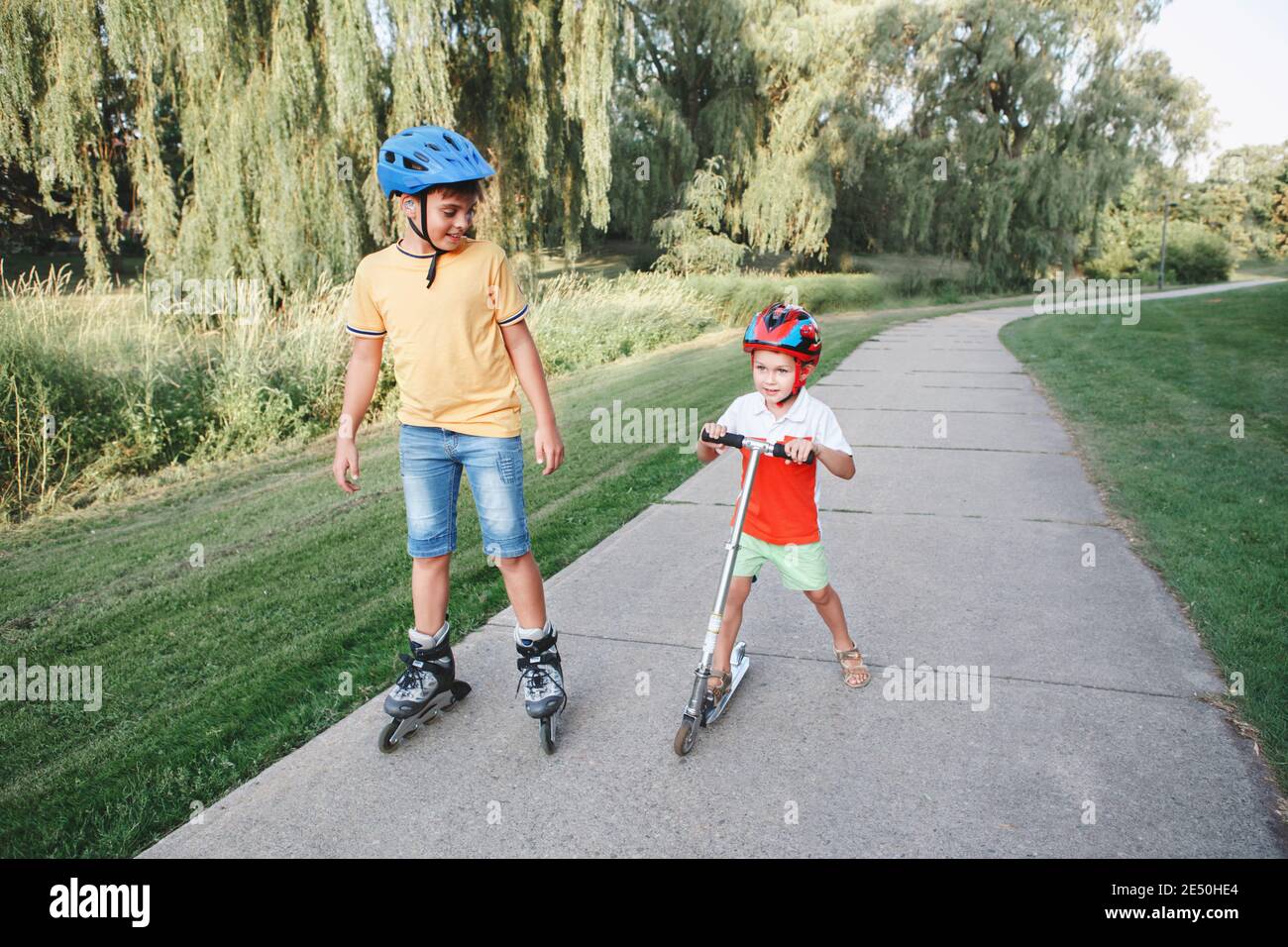 Ragazzi caucasici fratelli in caschi a cavallo pattini a rotelle e scooter su strada nel parco. Sport stagionale all'aperto per bambini. Infanzia sana Foto Stock