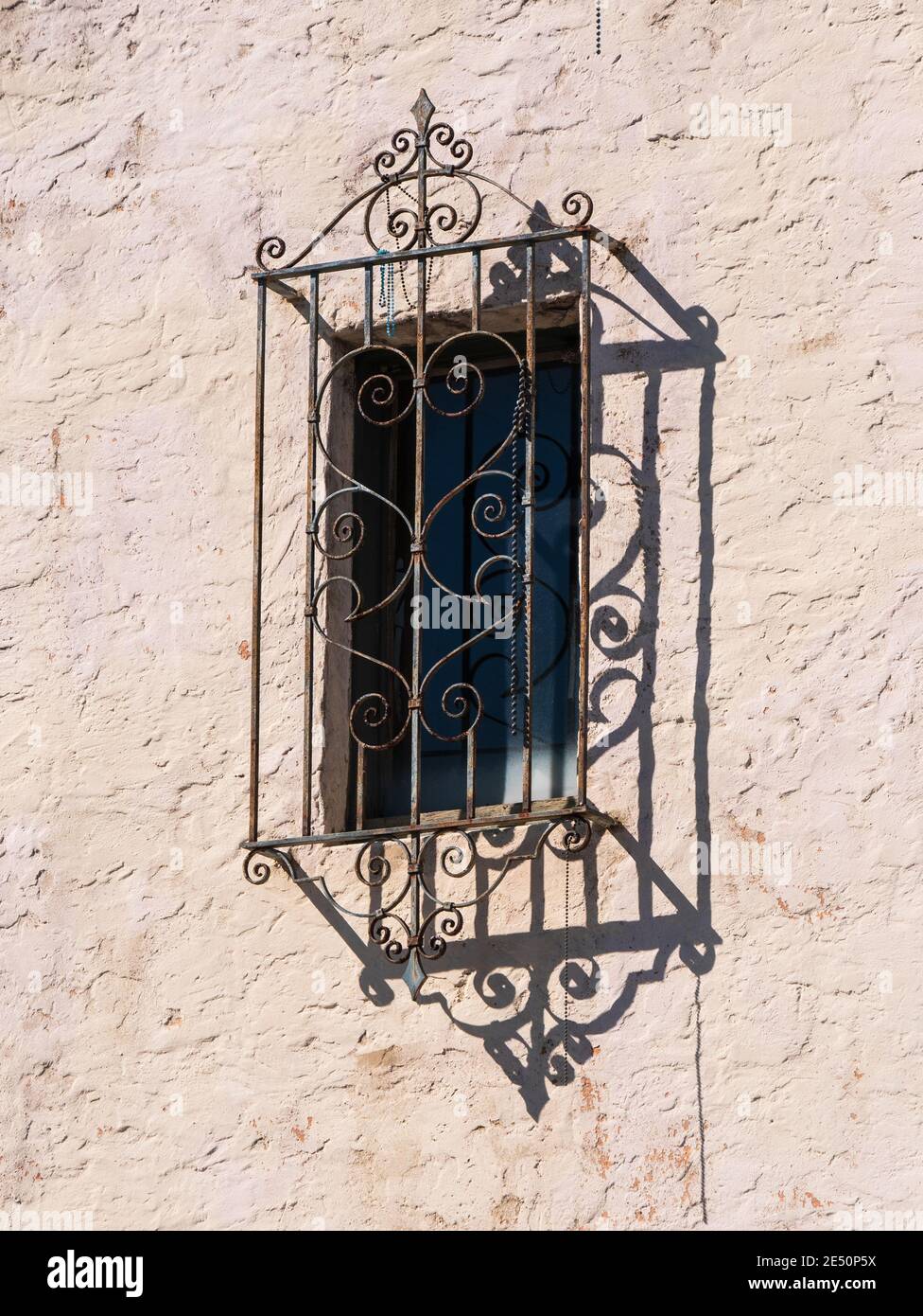 finestra con una protezione decorativa in ferro battuto sulla testurizzata parete in stucco di un edificio con forte ombra da luminosa luce solare Foto Stock