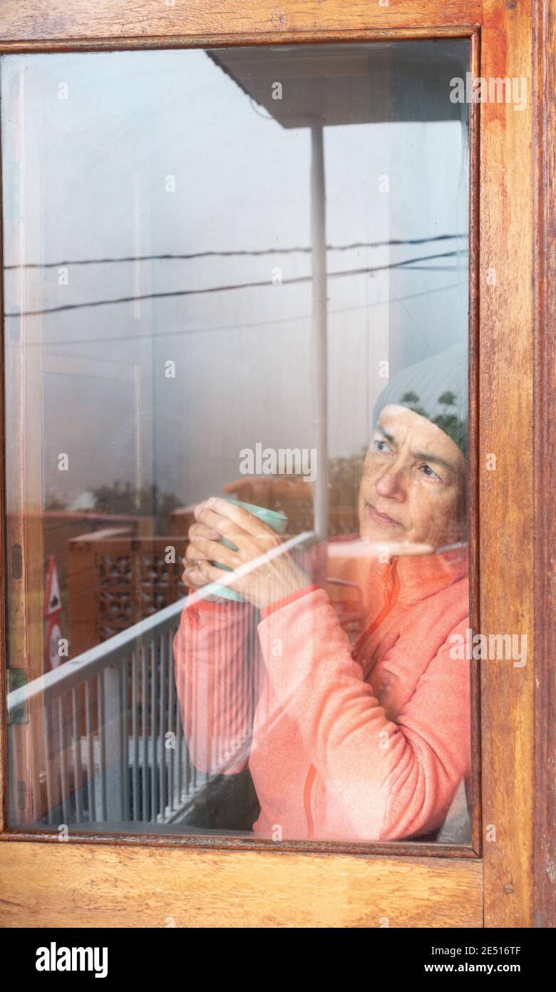 Donna di mezza età con una tazza di caffè che guarda fuori dalla finestra in una giornata fredda. Foto Stock