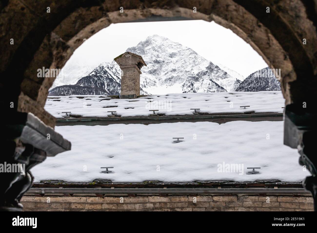 Un tetto innevato con un camino sulla cima e vette di montagna lontane, come si vede dall'arco rotondo di un chiostro Foto Stock