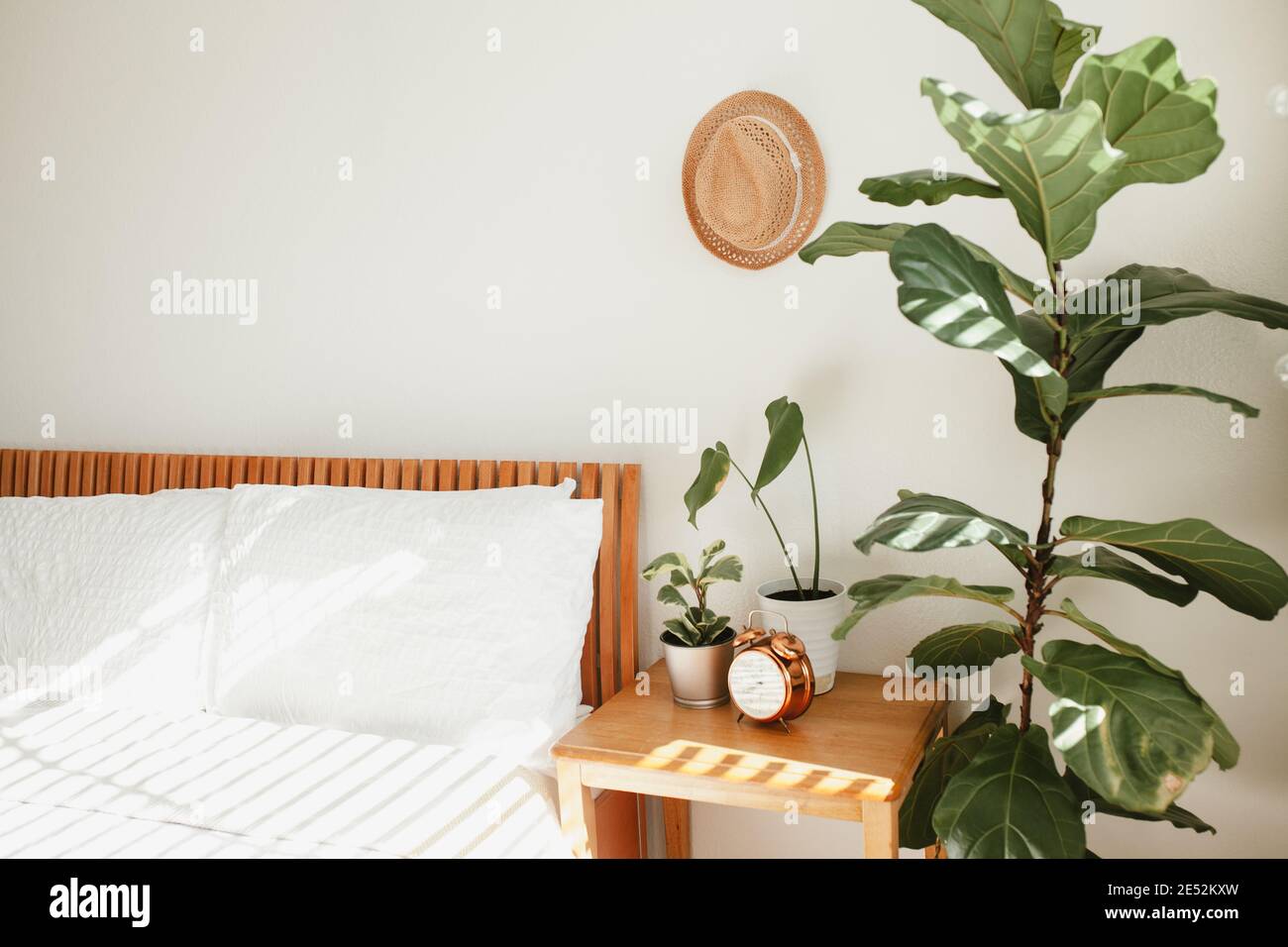 camera da letto bianca con letto in legno arancione, biancheria da letto bianca, comodino e piante vicino al letto, cappello di paglia sulla parete e orologio in rame vintage o Foto Stock