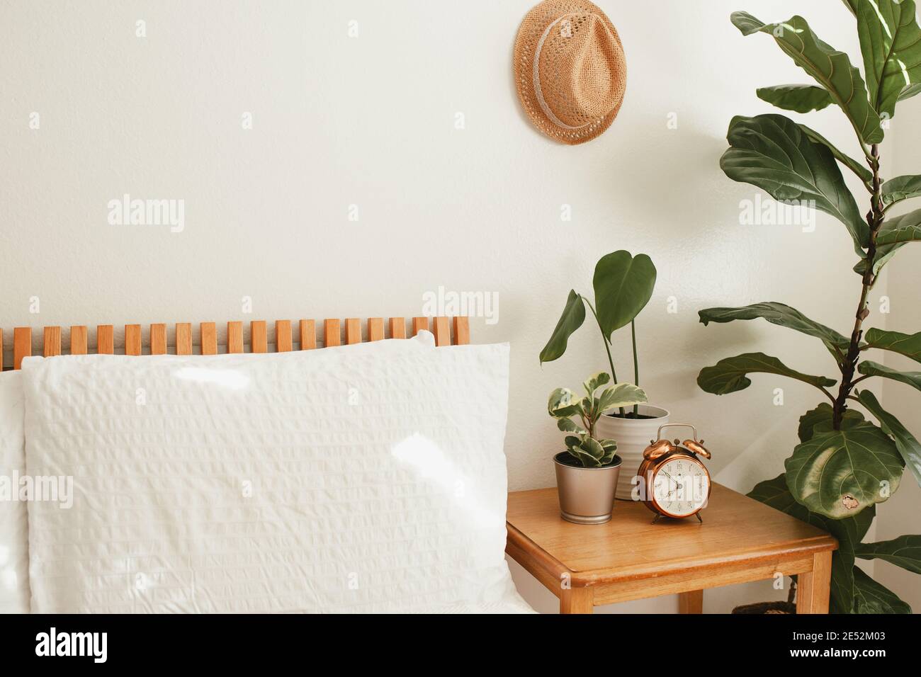 camera da letto bianca con letto in legno arancione, biancheria da letto bianca, comodino e piante vicino al letto, cappello di paglia sulla parete e orologio in rame vintage o Foto Stock