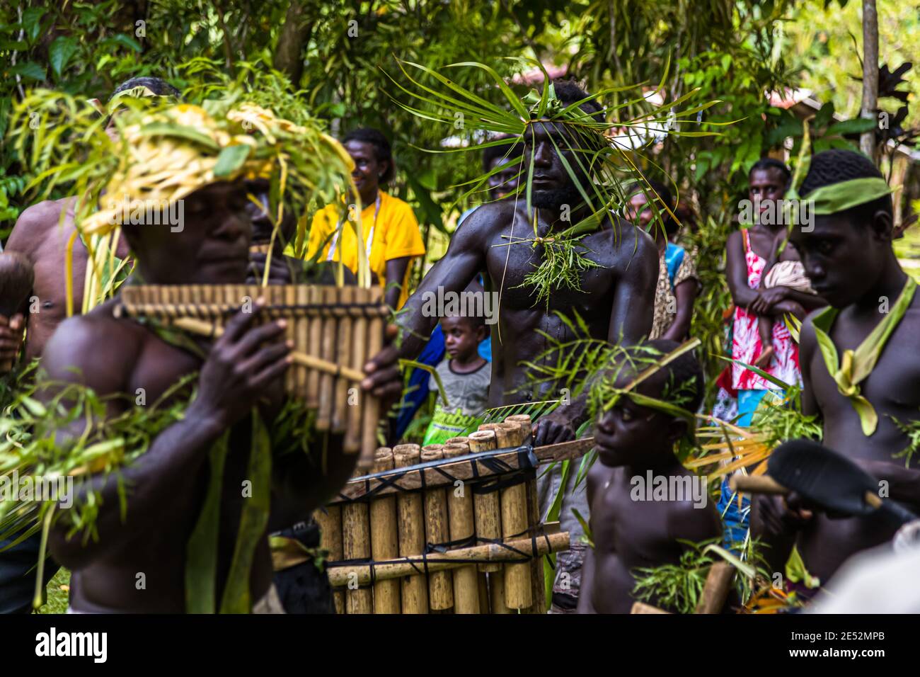 Un musicista che indossa un cappello intrecciato con foglie suona le panpipe durante un sing-sing tradizionale sull'isola di Tautsina, Bougainvillea, Papua Nuova Guinea, con la ballerina con copricapo di foglie verdi visibile dietro di lui. Foto Stock