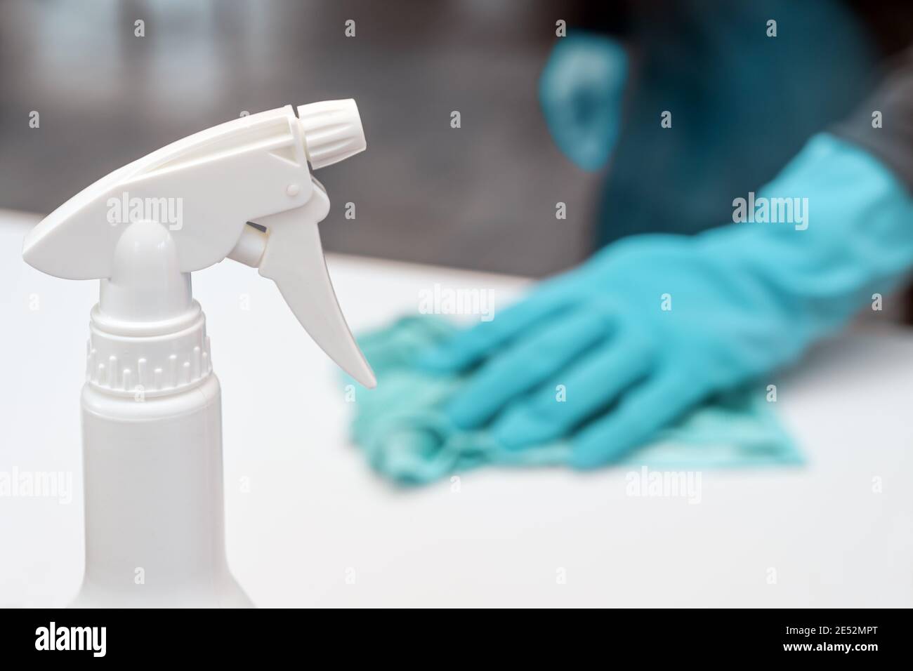 Flacone con spray disinfettante in primo piano, persona sfocata con guanti blu nella parte posteriore pulire la superficie bianca, per prevenire l'infezione da covid-19 Foto Stock