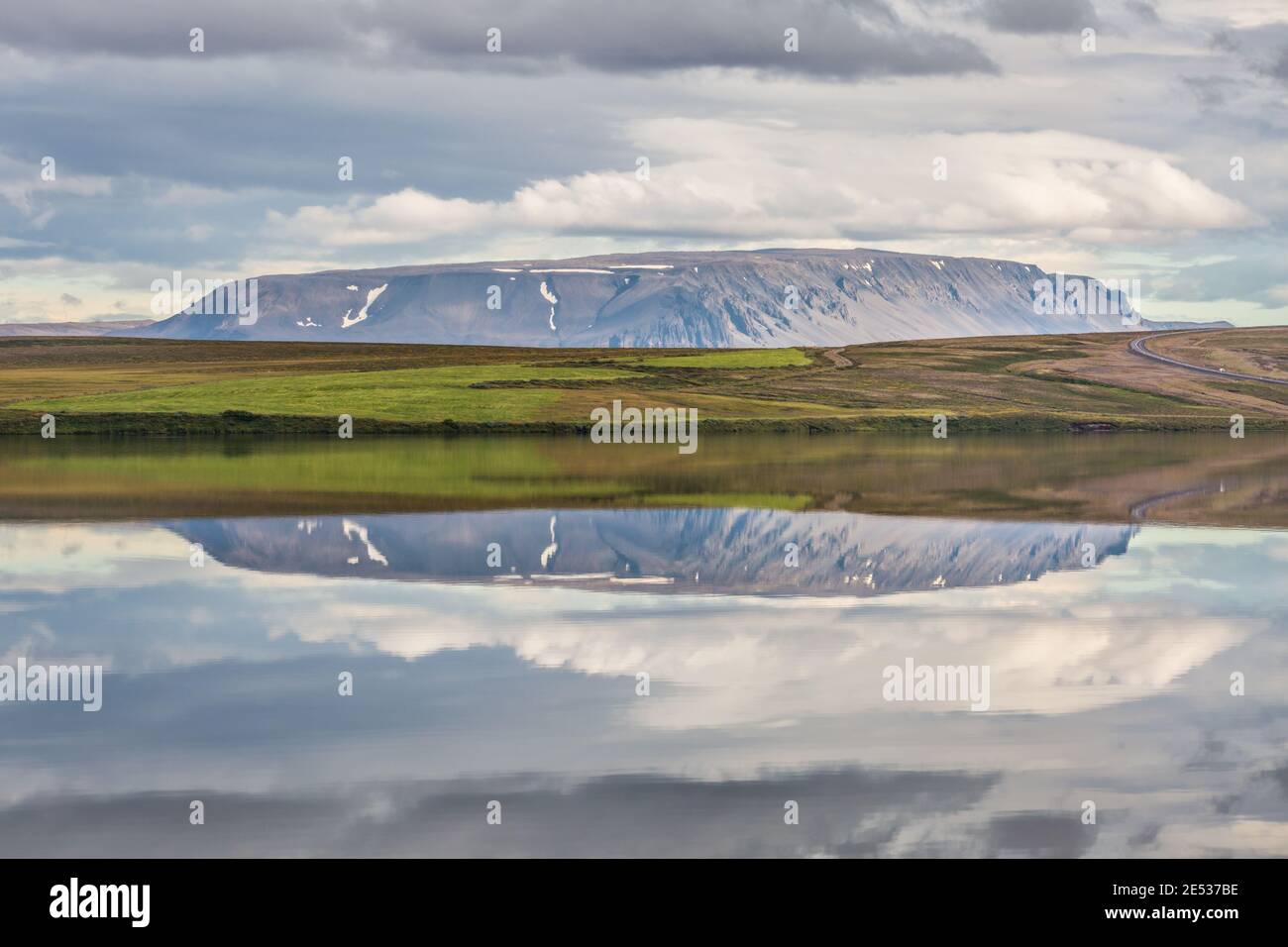 Paesaggio islandese con un prato e una montagna distante che riflette su acqua sotto un cielo nuvoloso Foto Stock
