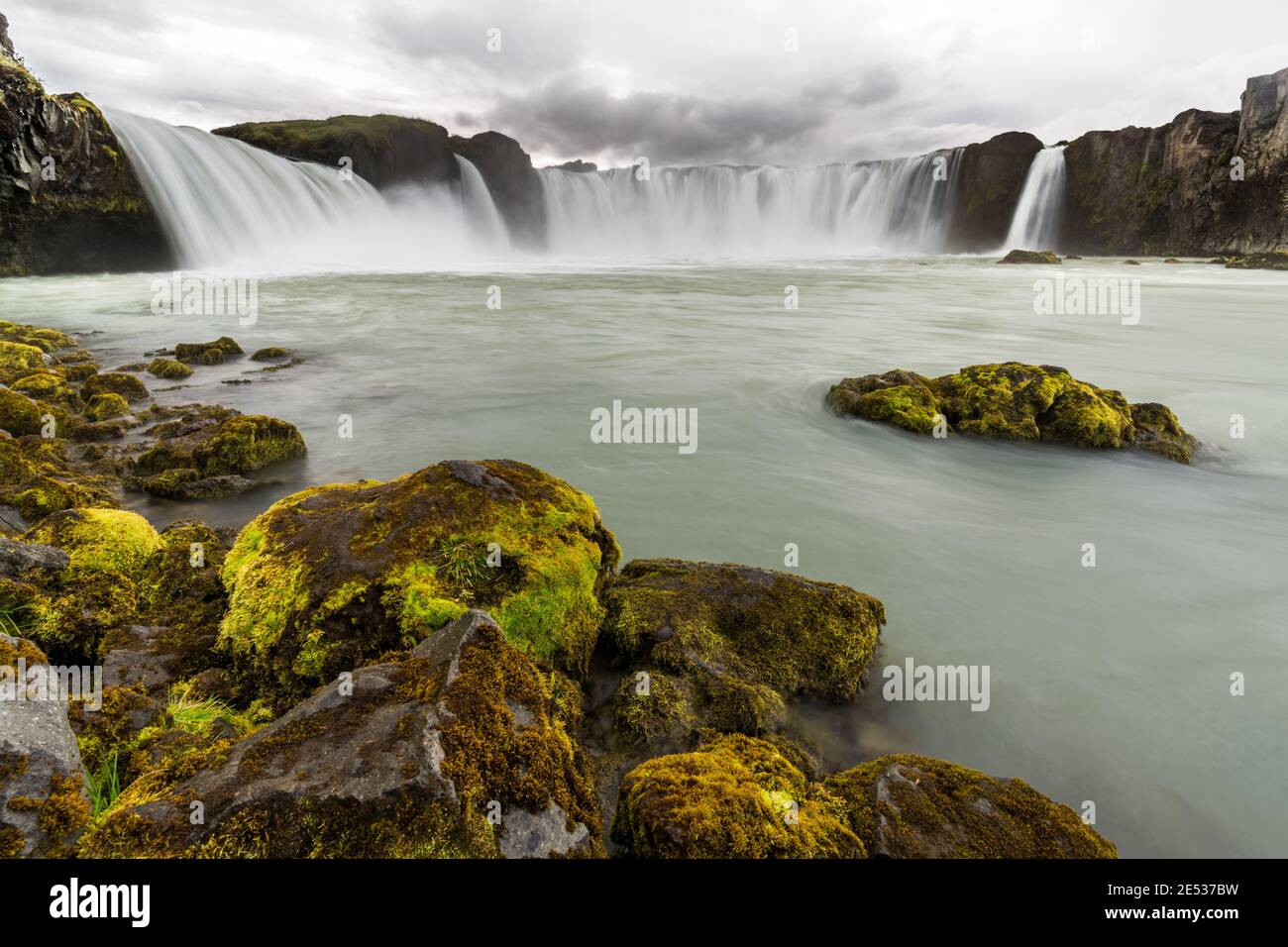 Paesaggio islandese con rocce ricoperte di muschio in primo piano e Godafoss, una delle grandi cascate, sullo sfondo, sotto un cielo nuvoloso Foto Stock