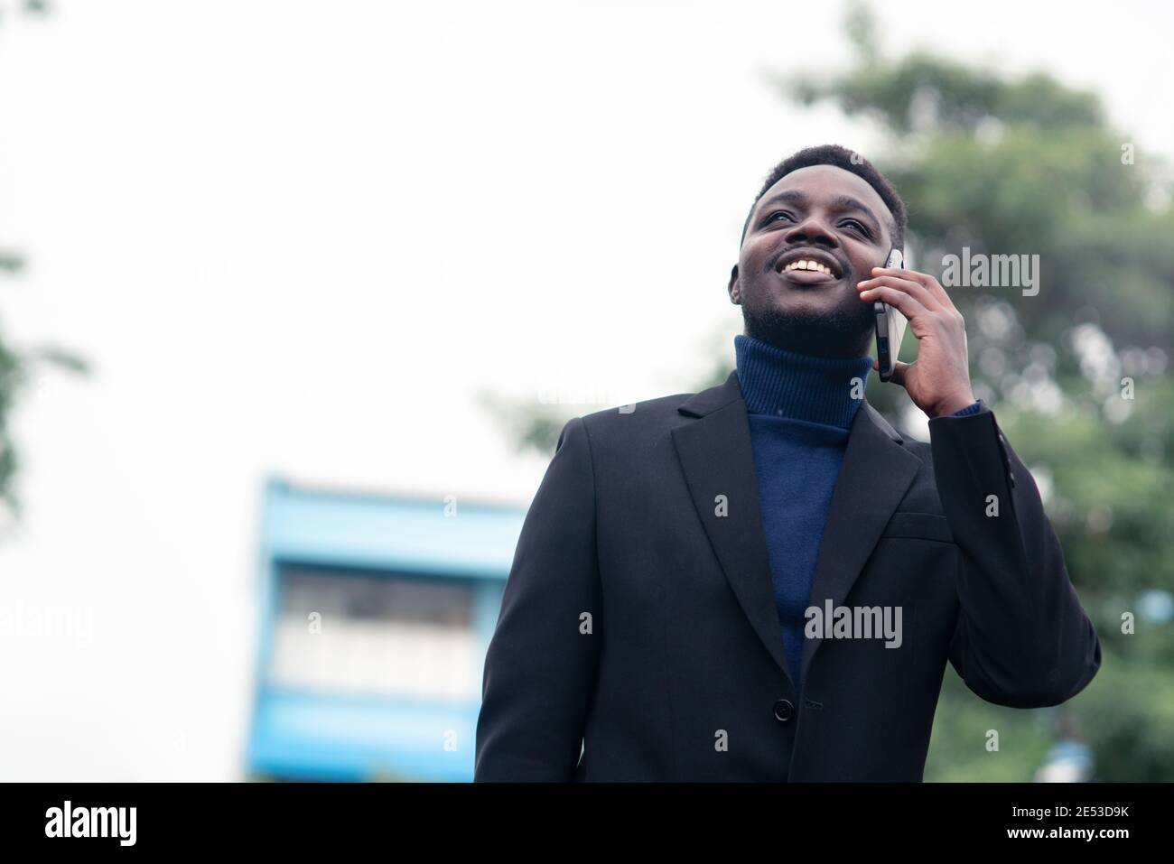 Bell'uomo d'affari africano che usa lo smartphone in un abito nero formale alla moda. Ragazzo con barba che indossa una manica lunga blu o un maglione Foto Stock