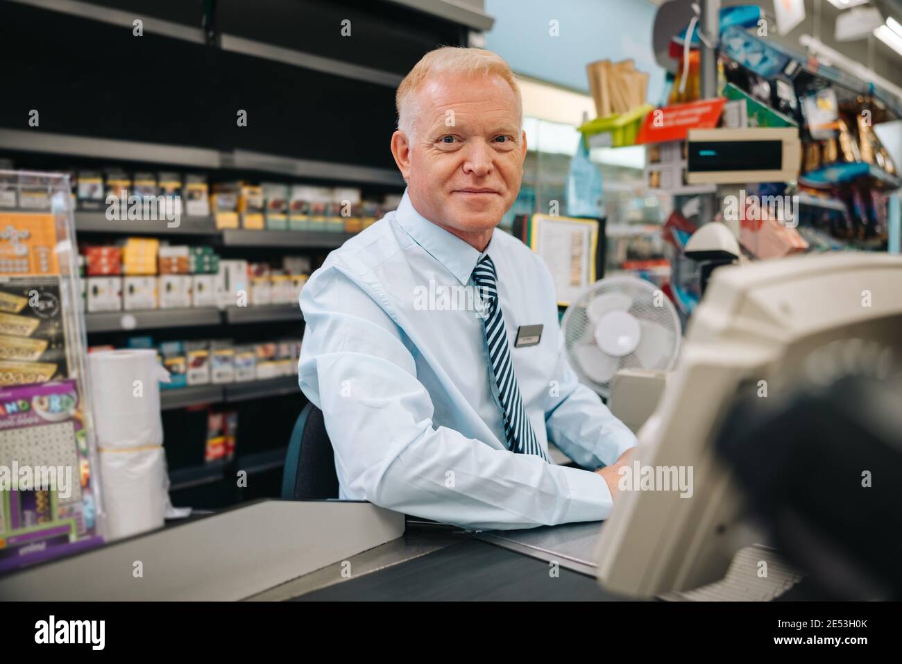 Ritratto di un uomo anziano che lavora alla cassa di super mercato e guarda la fotocamera. Negozio di alimentari cassiere al banco del check-out. Foto Stock