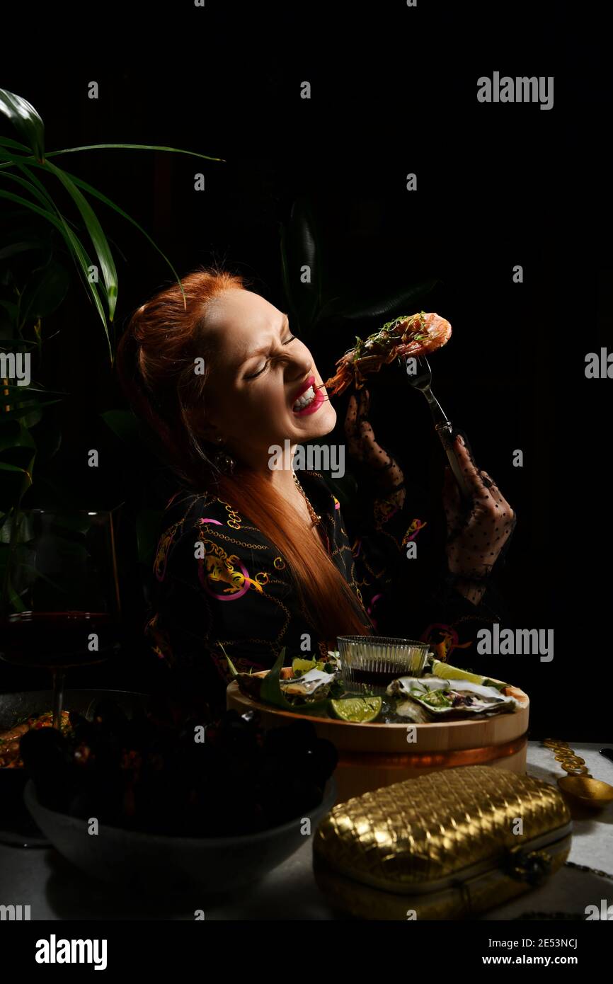 Una ricca donna dai capelli rossi che mangia nel ristorante di pesce tiene un gamberetto a forcella o una langoutine. Tavolo da pranzo al tavolo che serve ostriche fresche Foto Stock