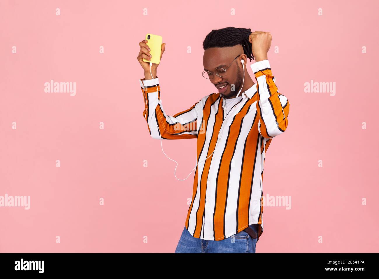 Uomo afro-americano positivo con dreadlock in occhiali e elegante a righe camicia che ascolta musica o radio sullo smartphone indossando l'auricolare e danci Foto Stock