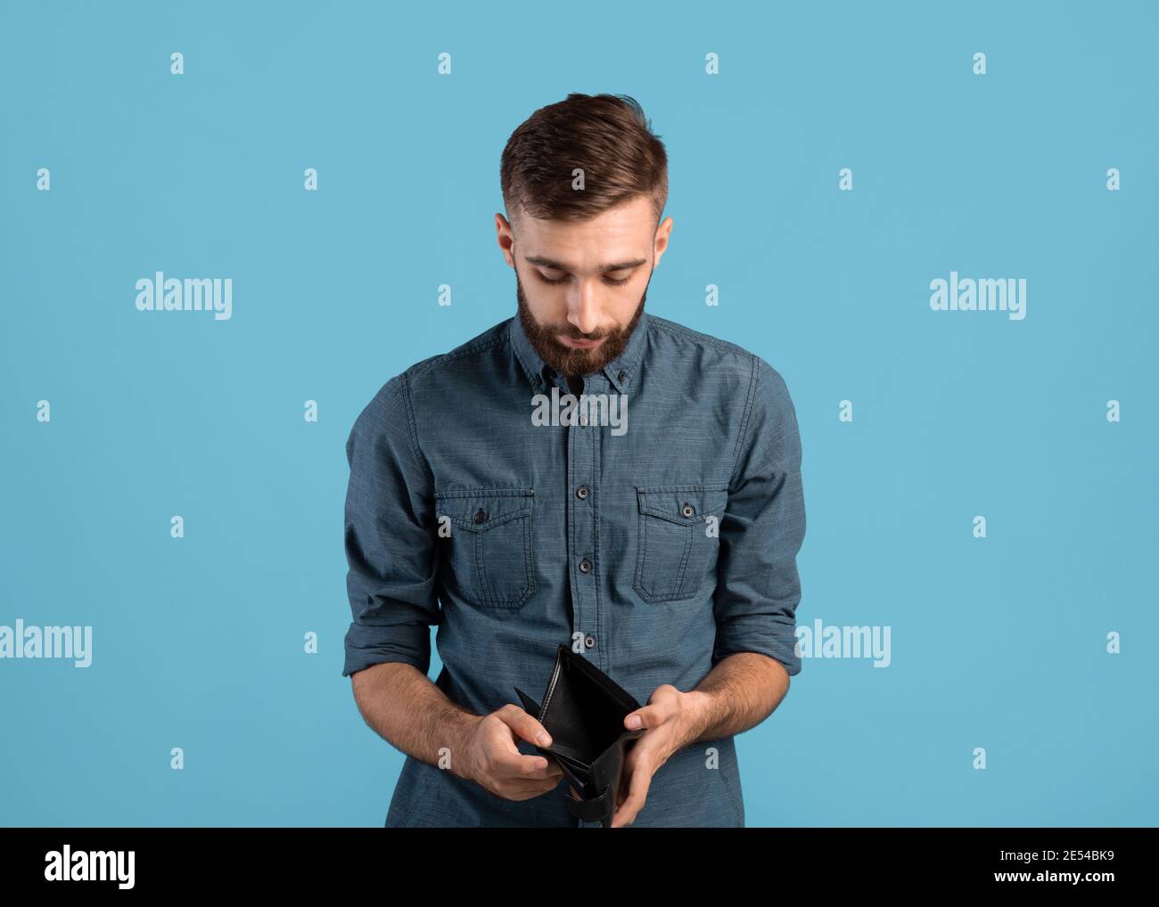 Povertà e disoccupazione. Disperato giovane uomo che mostra portafoglio vuoto senza soldi su sfondo blu studio Foto Stock