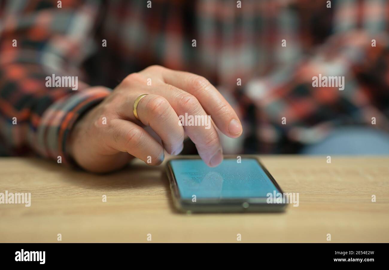 Un uomo sta leggendo le notizie sul suo telefono cellulare, che si trova su un tavolo di legno. Primo piano sull'anello d'oro sul dito destro di un uomo Foto Stock