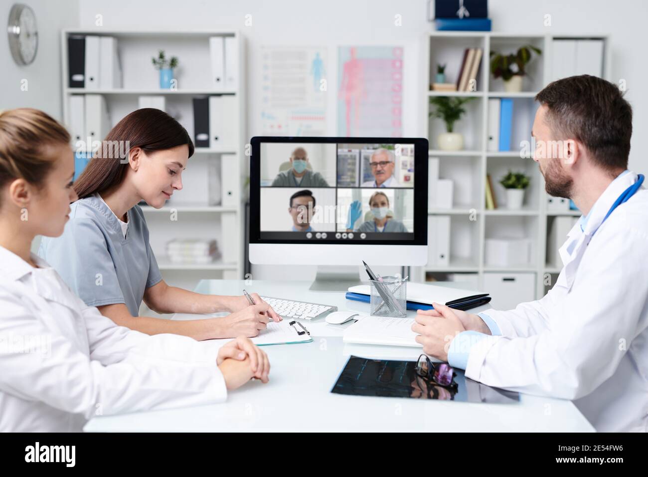 Gruppo di giovani professionisti che si consultano con i colleghi durante la comunicazione online mentre si è seduti davanti al monitor del computer in un ufficio medico Foto Stock