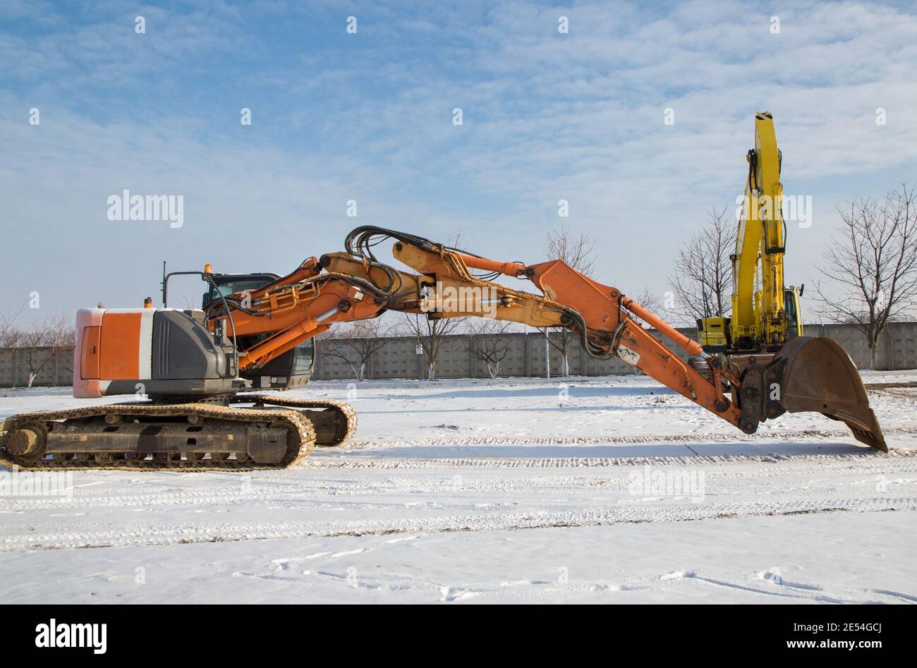 Due escavatori cingolati di grandi dimensioni sono in piedi in un cantiere. Un potente escavatore arancione in primo piano con una freccia estesa sopra la neve-c. Foto Stock