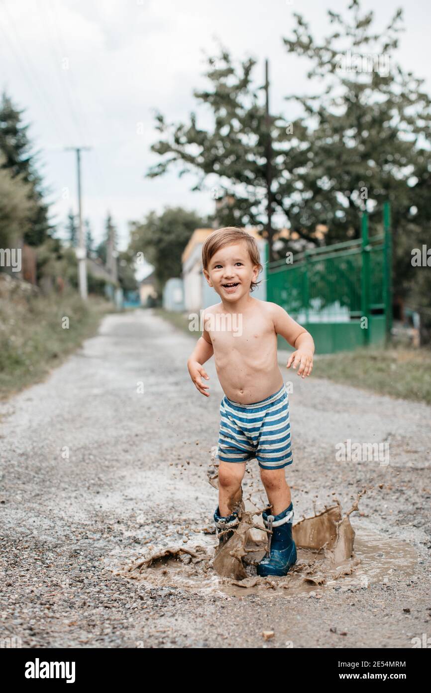 Ritratto di un bambino sorridente in stivali da pioggia che saltano le pudddle in una calda giornata estiva. Felice bambino in stivali wellington con divertimento spruzzi aroun Foto Stock