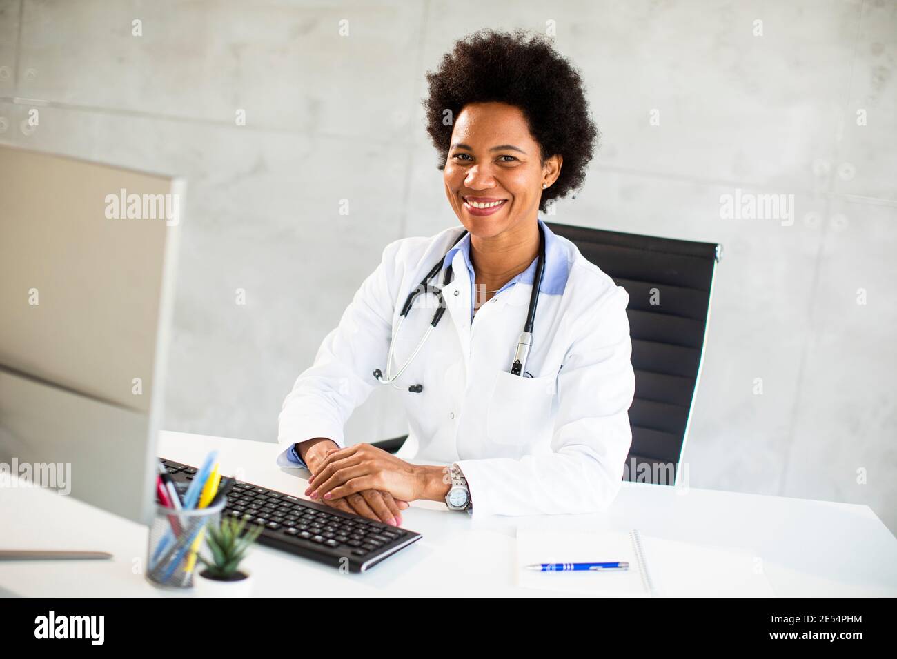 Donna Africana americana medico indossando camice bianco con stetoscopio seduta dietro la scrivania in ufficio Foto Stock