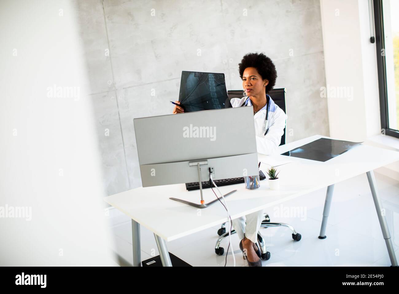 Donna Africana americana medico indossando camice bianco con stetoscopio seduta dietro la scrivania in ufficio e guardando l'immagine radiografica Foto Stock