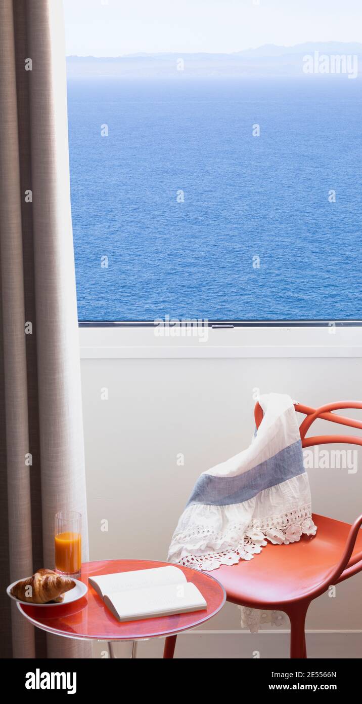 Finestra con vista sul mare, tavolino da caffè rosso e sedia con prima colazione a base di croissant e succo di frutta, libro e abito estivo Foto Stock