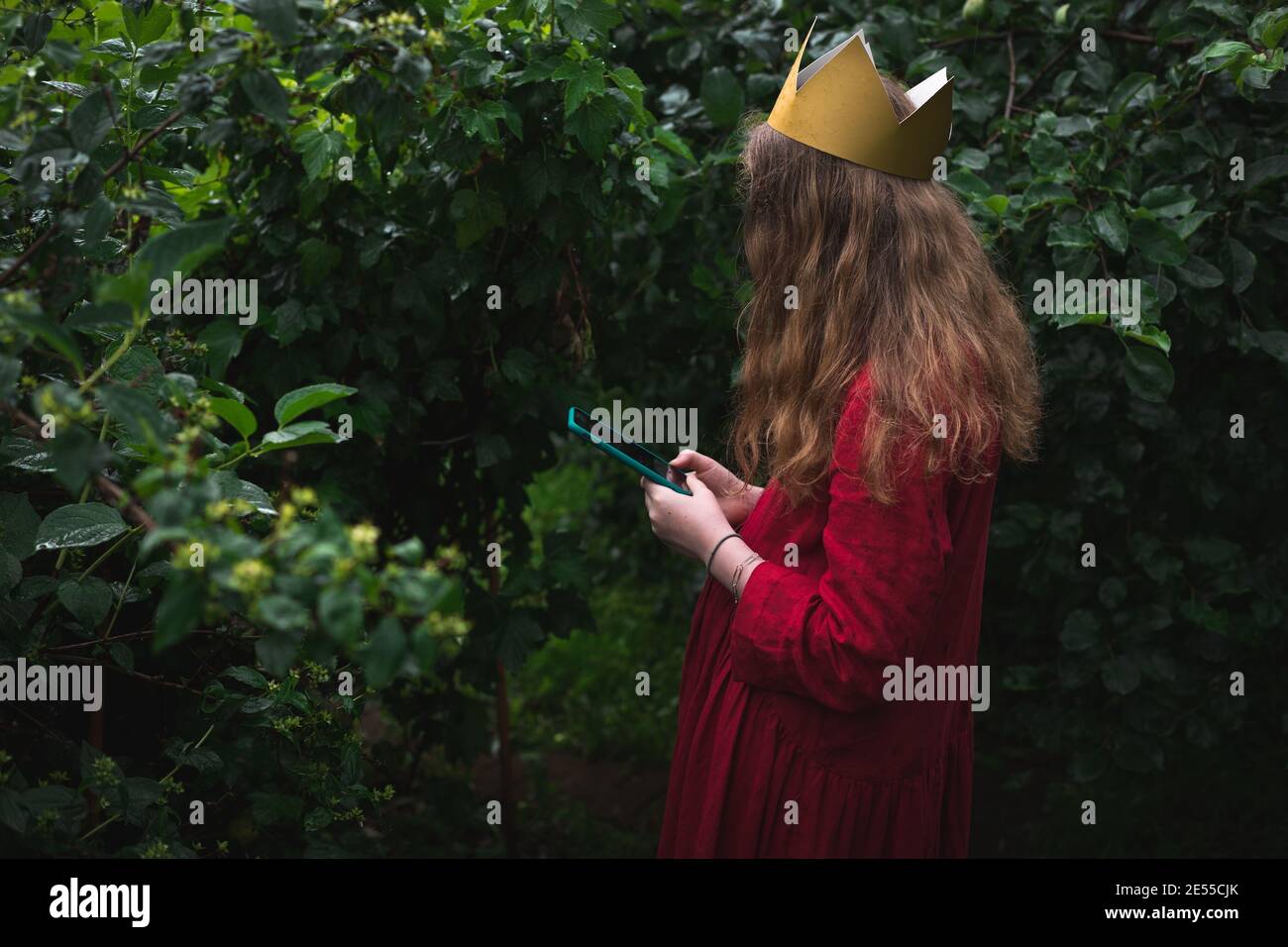 Pre ragazza indossando vestito rosso e corona di carta in piedi giardino bagnato utilizzando il telefono Foto Stock