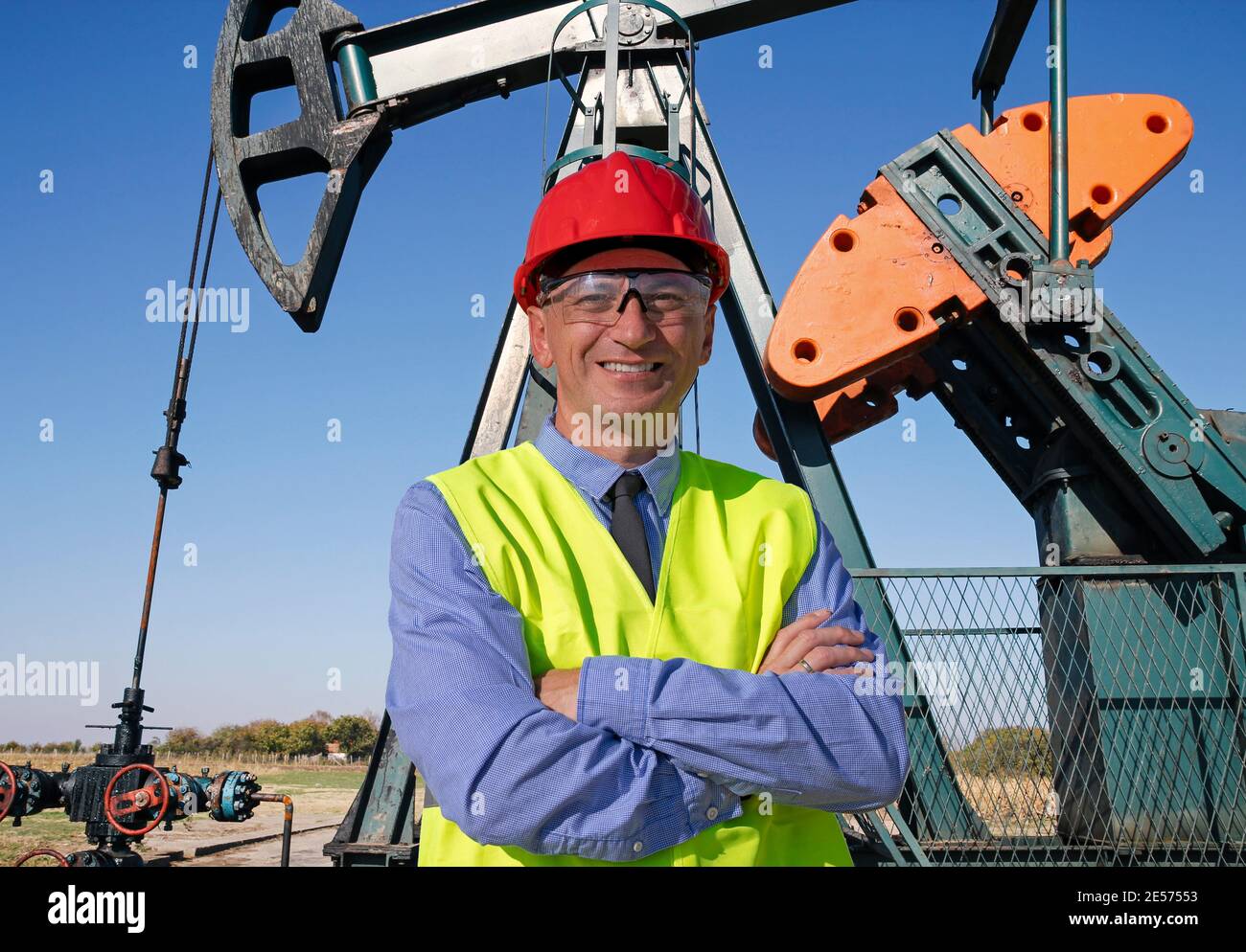 Ingegnere del petrolio e del gas in Red Hardhat e Yellow West in piedi davanti al martinetto della pompa del pozzo del petrolio. Felice uomo d'affari che guarda la fotocamera. Foto Stock