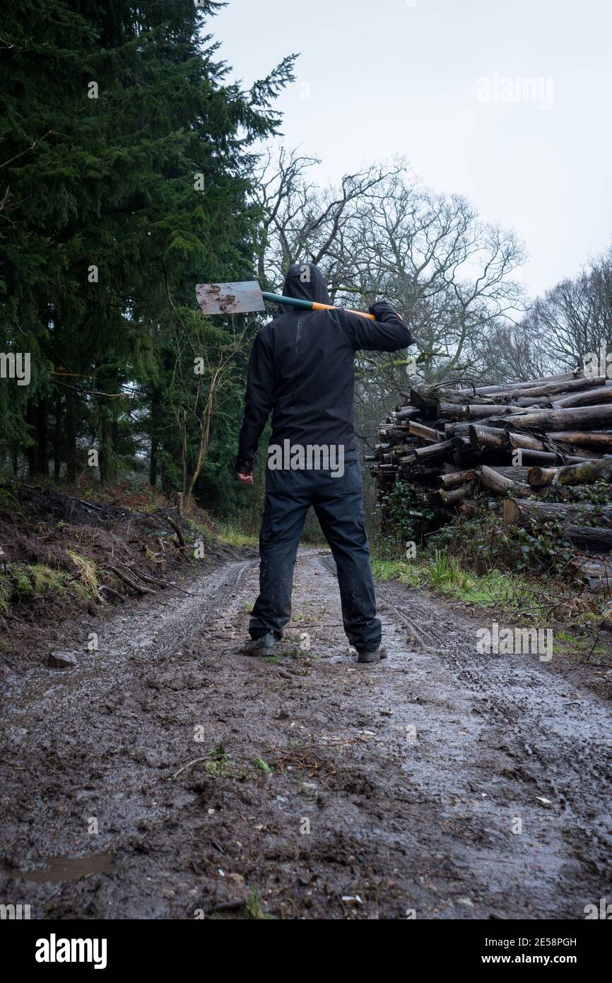 Un uomo incappucciato, di nuovo alla macchina fotografica su un sentiero fangoso che tiene una vanga, accanto a un mucchio di tronchi in una giornata di luna grigia in campagna Foto Stock