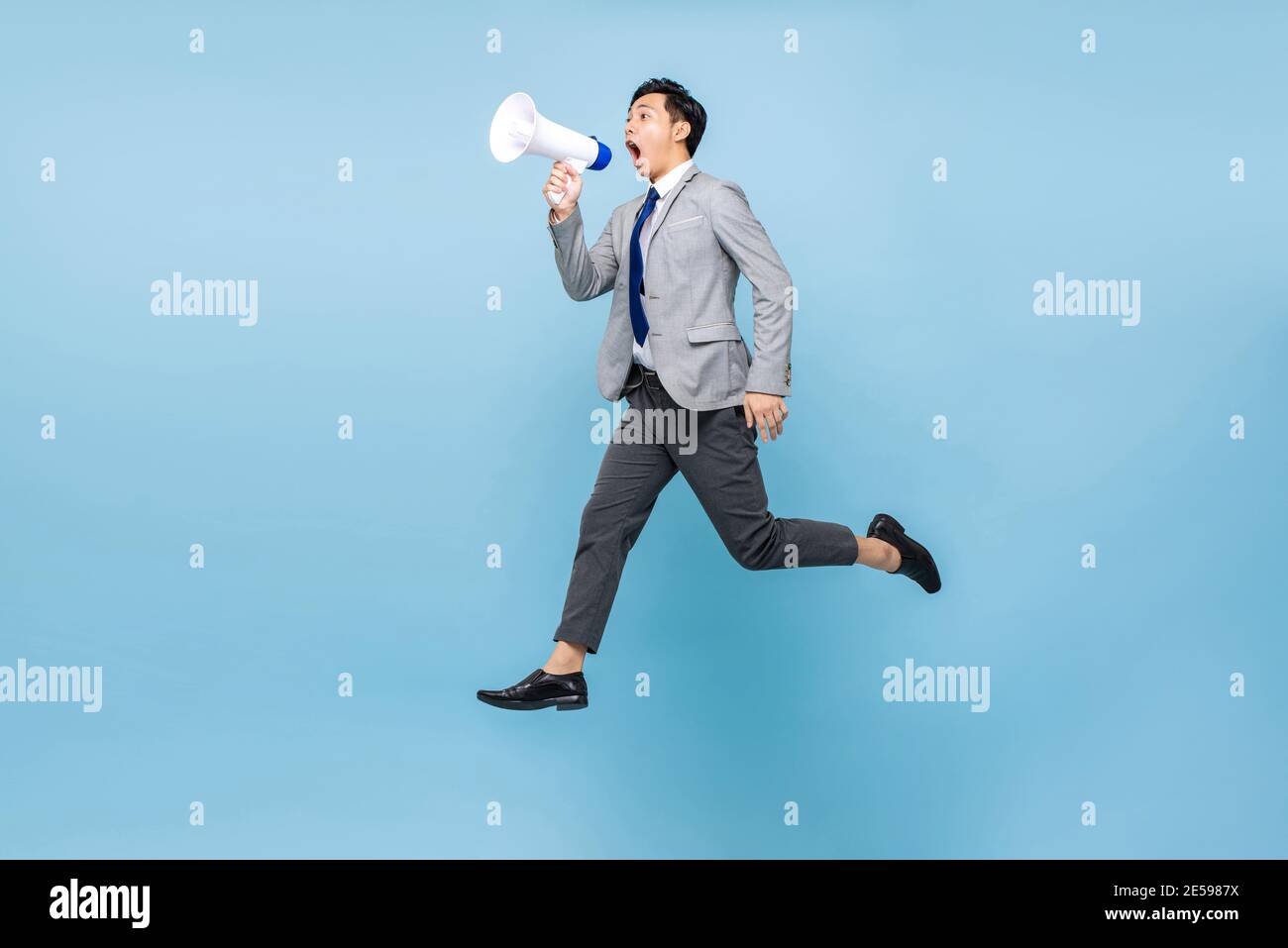 Giovane uomo d'affari asiatico che salta e grida sul megafono isolato sopra sfondo di colore blu chiaro Foto Stock