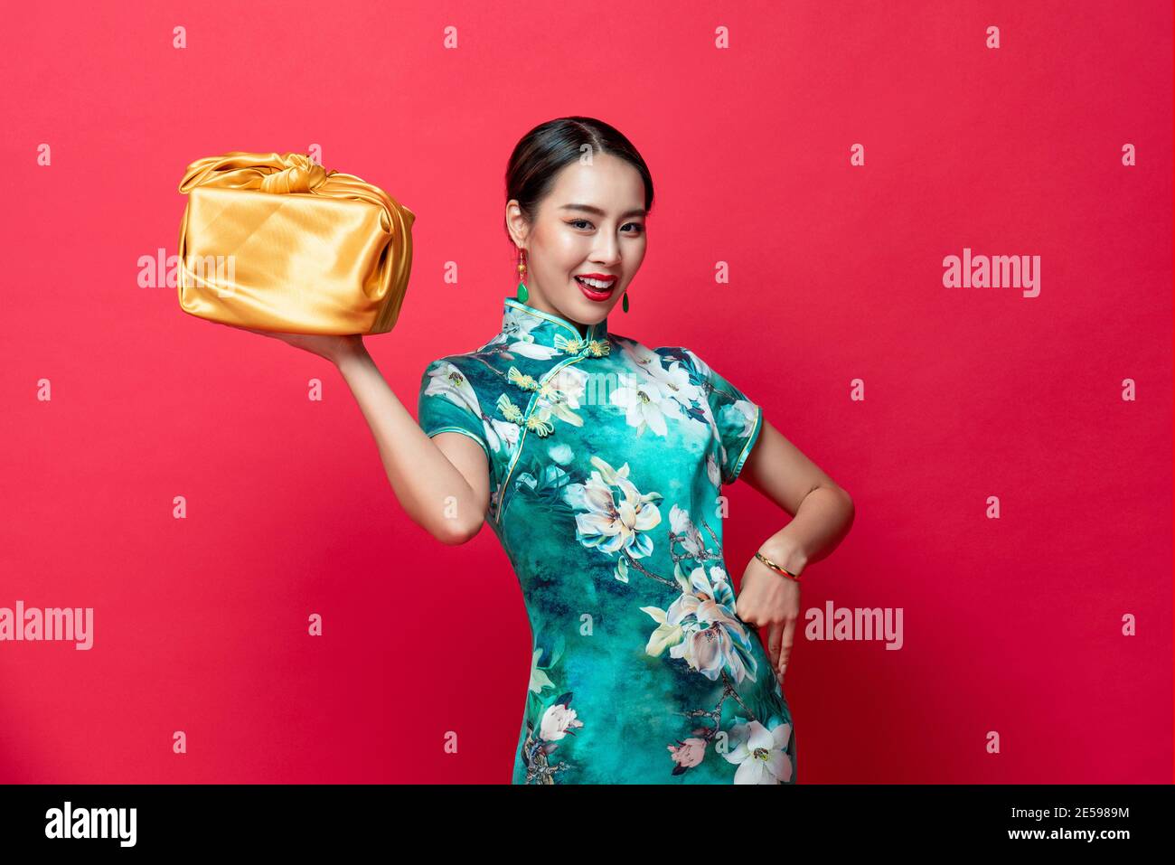 Sorridente bella donna asiatica in abito tradizionale che tiene dono d'oro Box in rosso isolato sfondo studio per il cinese nuovo anno concetti Foto Stock