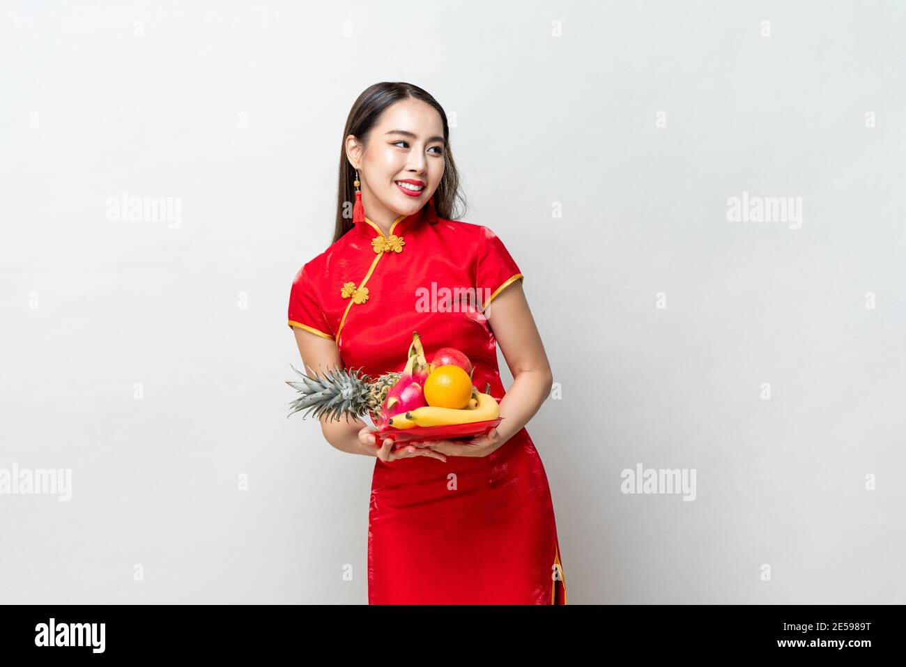 Giovane donna asiatica in abito traditinal rosso cheongsam tenendo fortunato Frutta per il nuovo anno cinese in uno studio isolato grigio chiaro sfondo Foto Stock