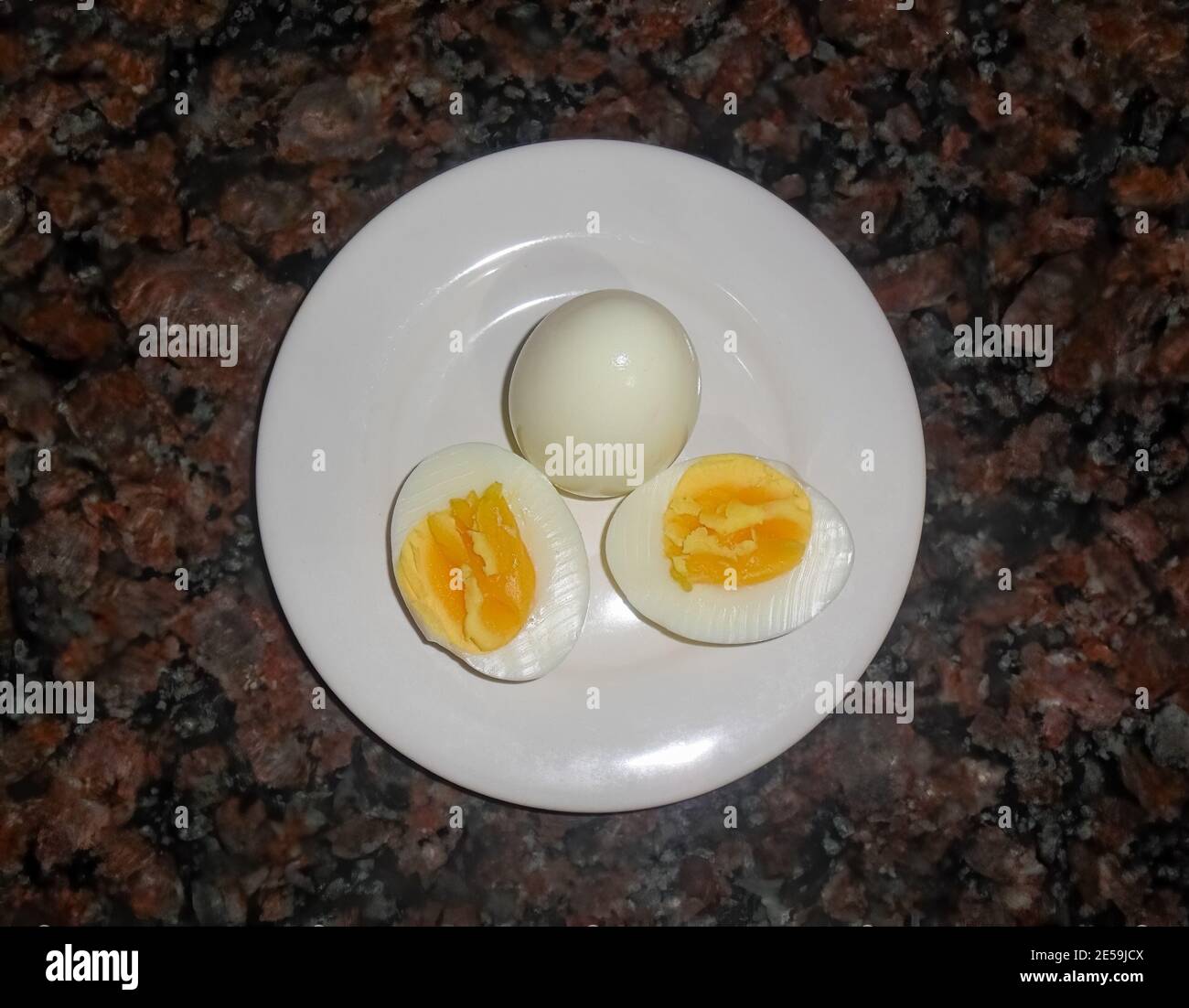 Due uova bollite su un piatto bianco, una a metà. Mostrando tuorlo. Sfondo di granito scuro. Overhead Foto Stock