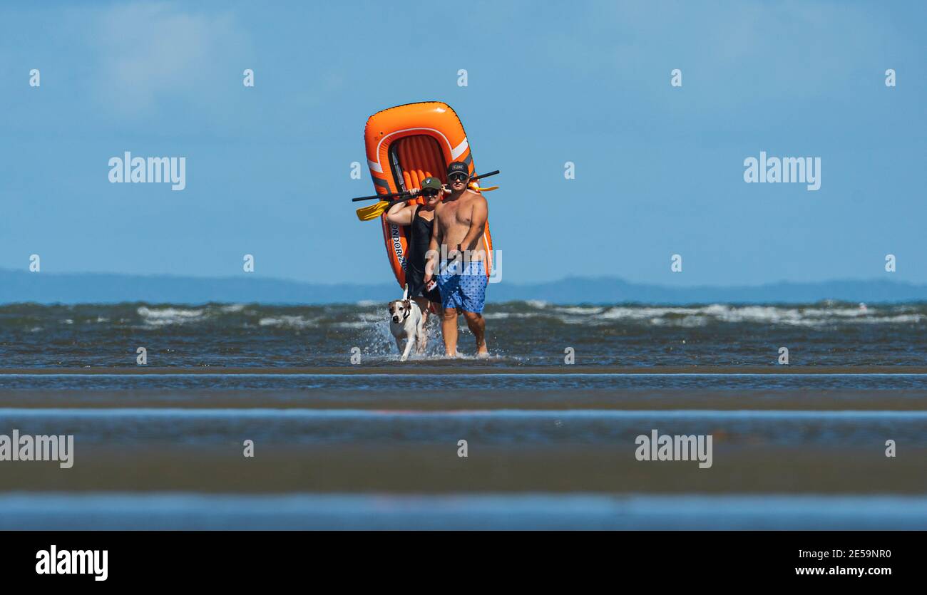 Coppia che porta una barca di gomma e che cammina un cane sulla spiaggia a bassa marea a Beachmere, Queensland, QLD, Australia Foto Stock