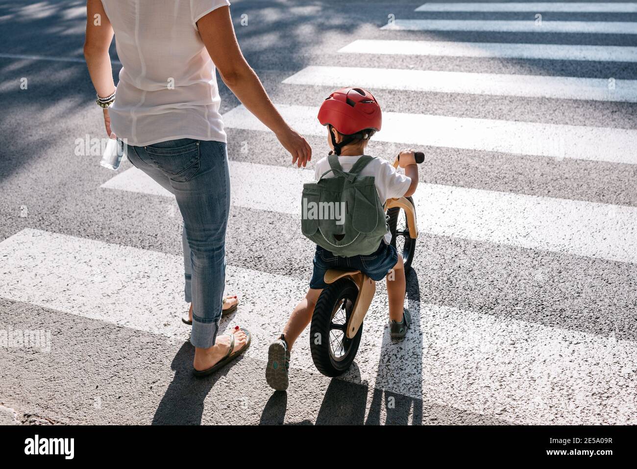 Vista posteriore di un bambino su una bici di equilibrio che attraversa una strada con la madre. Foto Stock