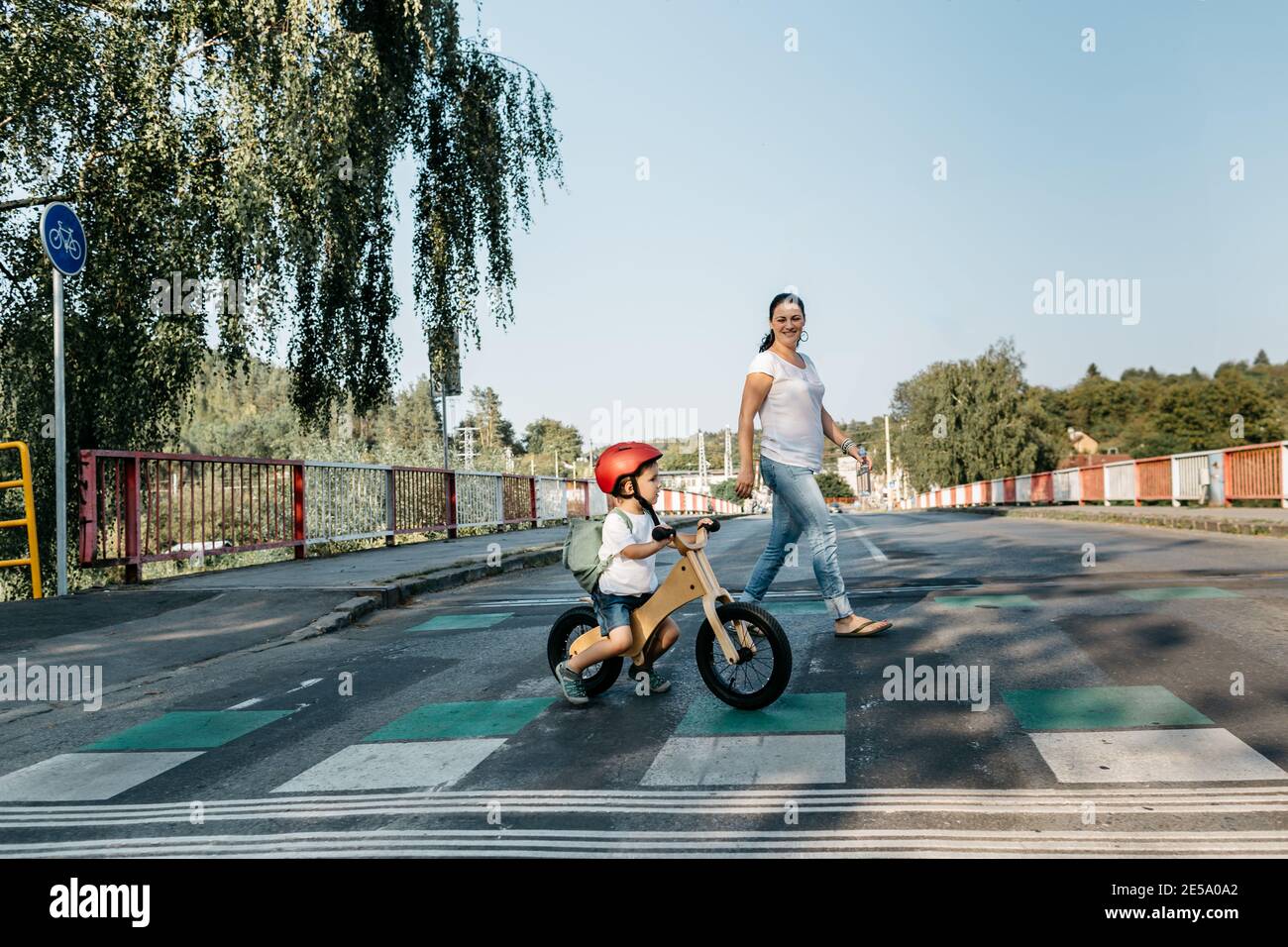 Vista laterale del bambino sulla bici di equilibrio e sua madre che attraversa la strada. Foto Stock