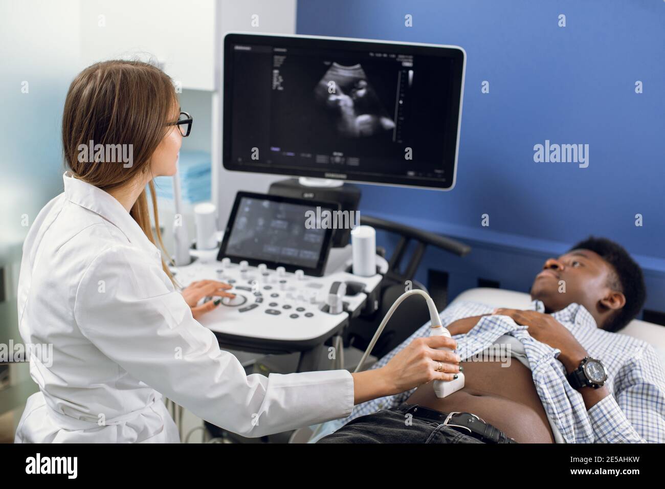 Giovane donna caucasica dottore che sposta trasduttore a ultrasuoni sulla pancia dell'uomo africano in ospedale. Dottore femmina che fa ultra suono di organi interni di Foto Stock