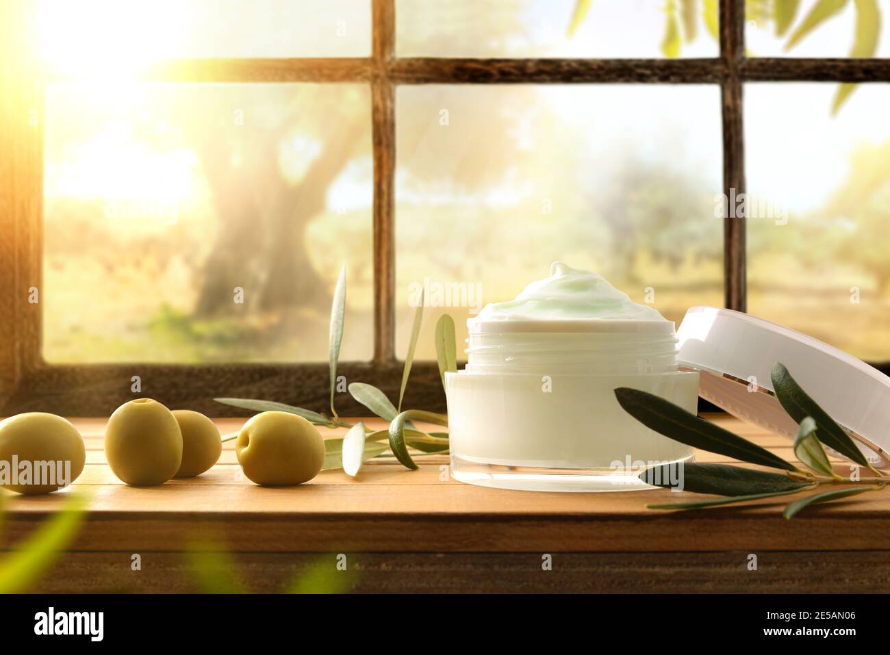 Crema naturale di pelle con estratto di oliva con rametto e olive su tavola di legno con fondo finestra e campo d'oliva. Vista dall'alto. Foto Stock