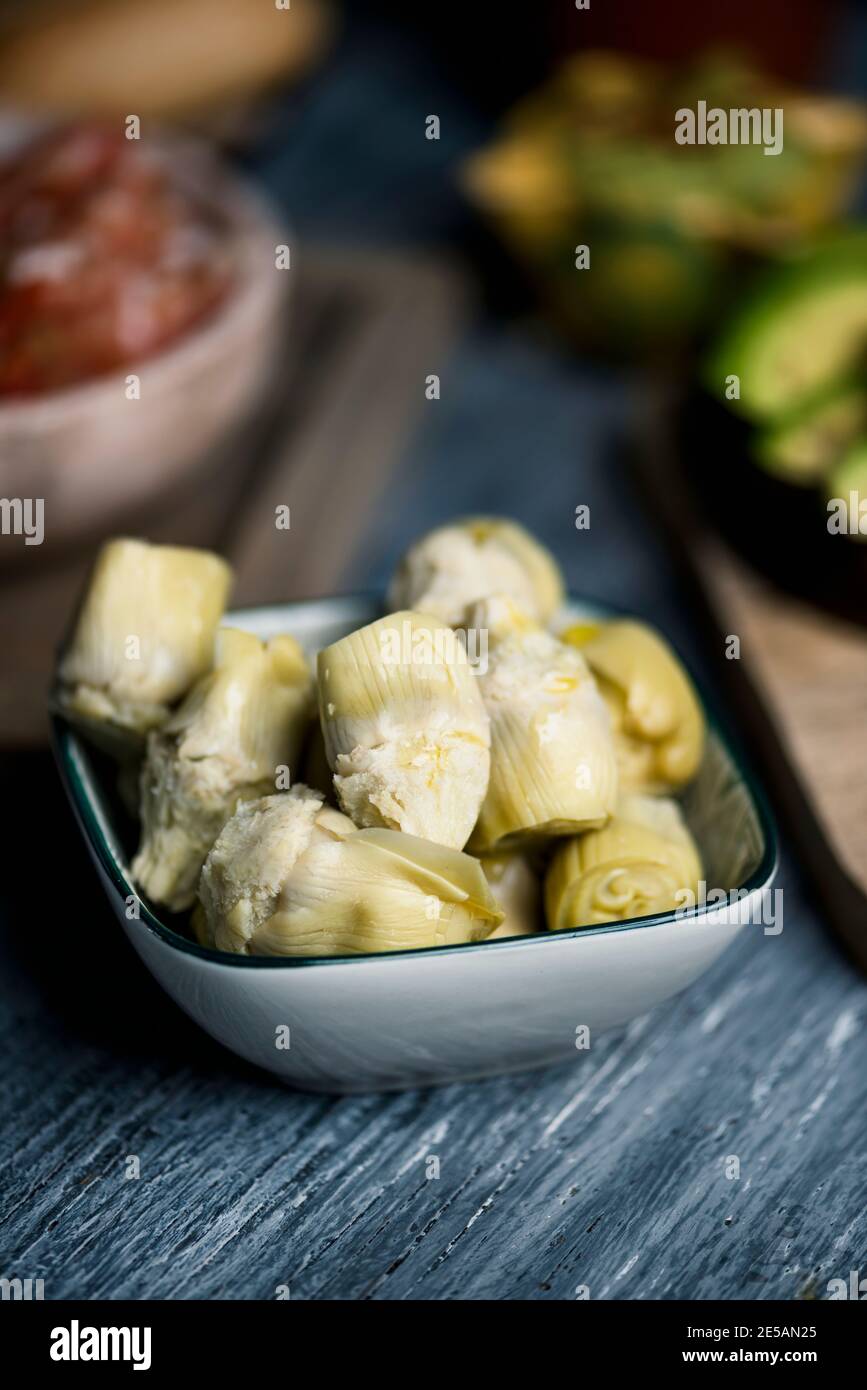 primo piano di una ciotola rettangolare in ceramica con alcuni cuori di carciofi cotti, su un tavolo grigio rustico in legno accanto ad alcuni altri ingredienti diversi da preparare Foto Stock