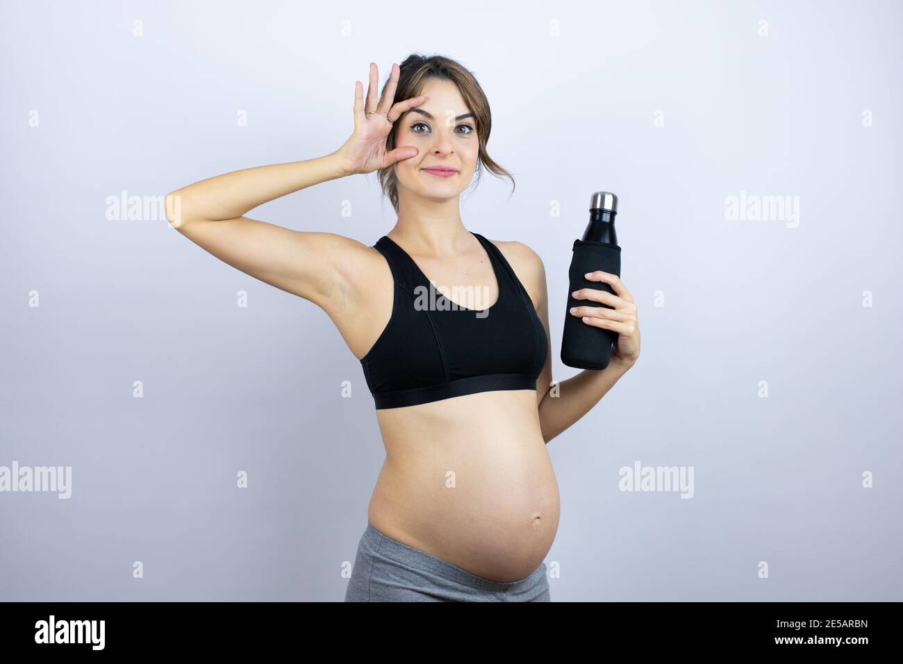 Giovane sportivo incinta indossando sportswear tenendo bottiglia con acqua su sfondo bianco cercando di aprire gli occhi con le dita, sonnolento e stanco per m Foto Stock